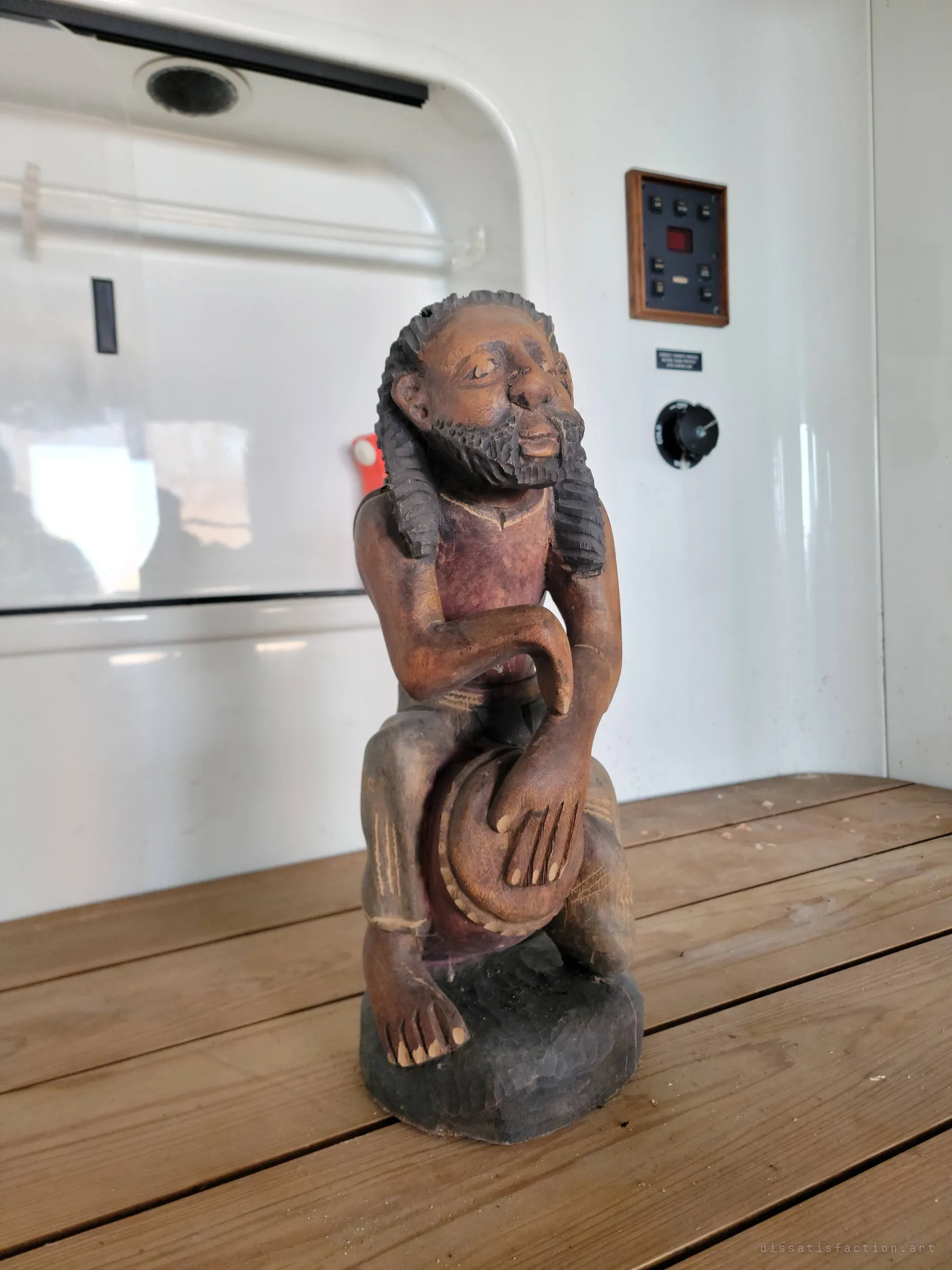 A hand-carved wooden figure of a bearded drummer sits on a slatted wooden bench against a white panelled wall with a small control panel. A strange, watchful ornament left behind in an empty room.