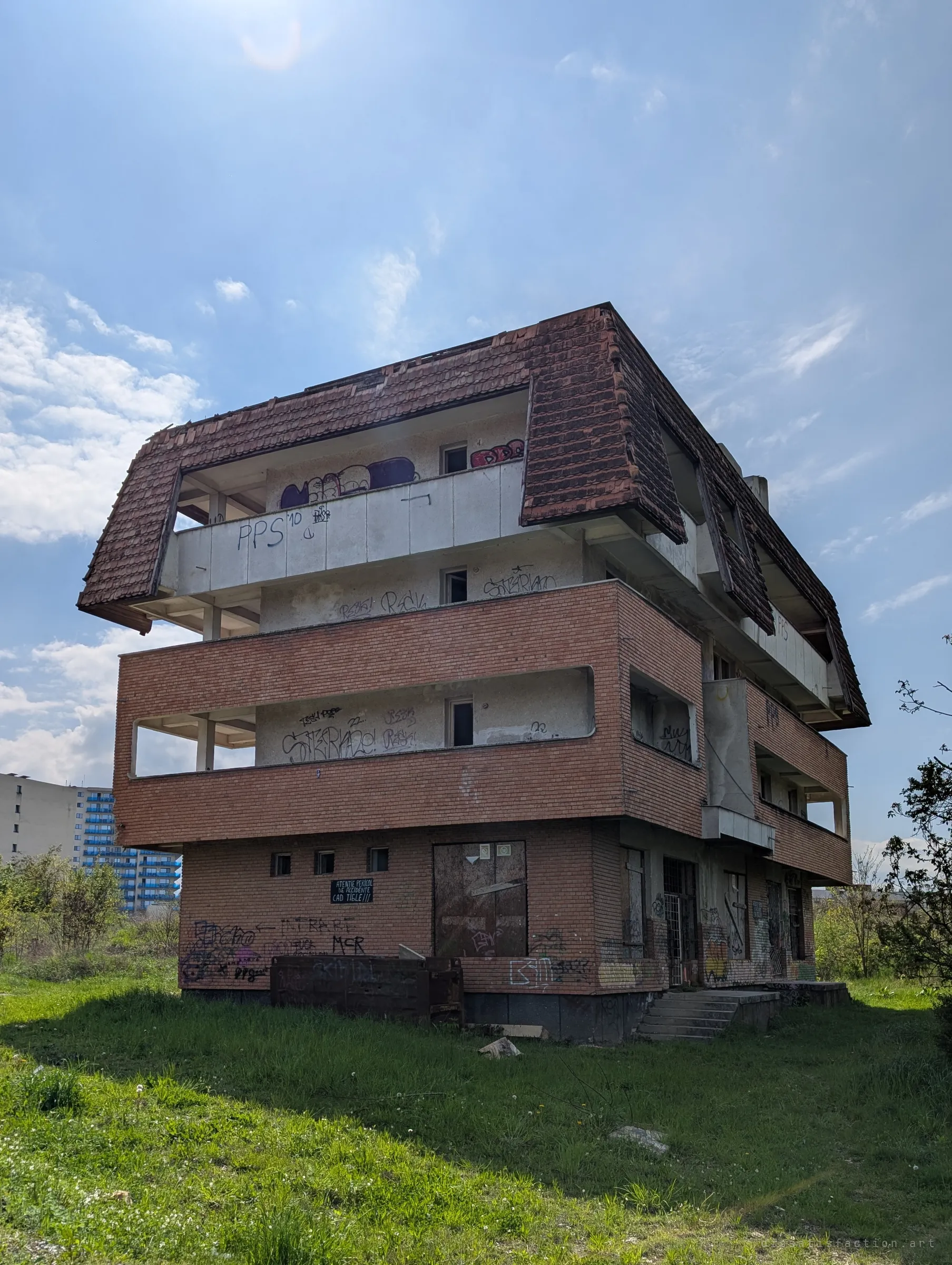 Low-angle view of an abandoned brick-and-concrete block under a sagging tiled mansard roof, its open balconies covered in graffiti. It sets the scene: a mid-rise building left to rot in an overgrown lot.