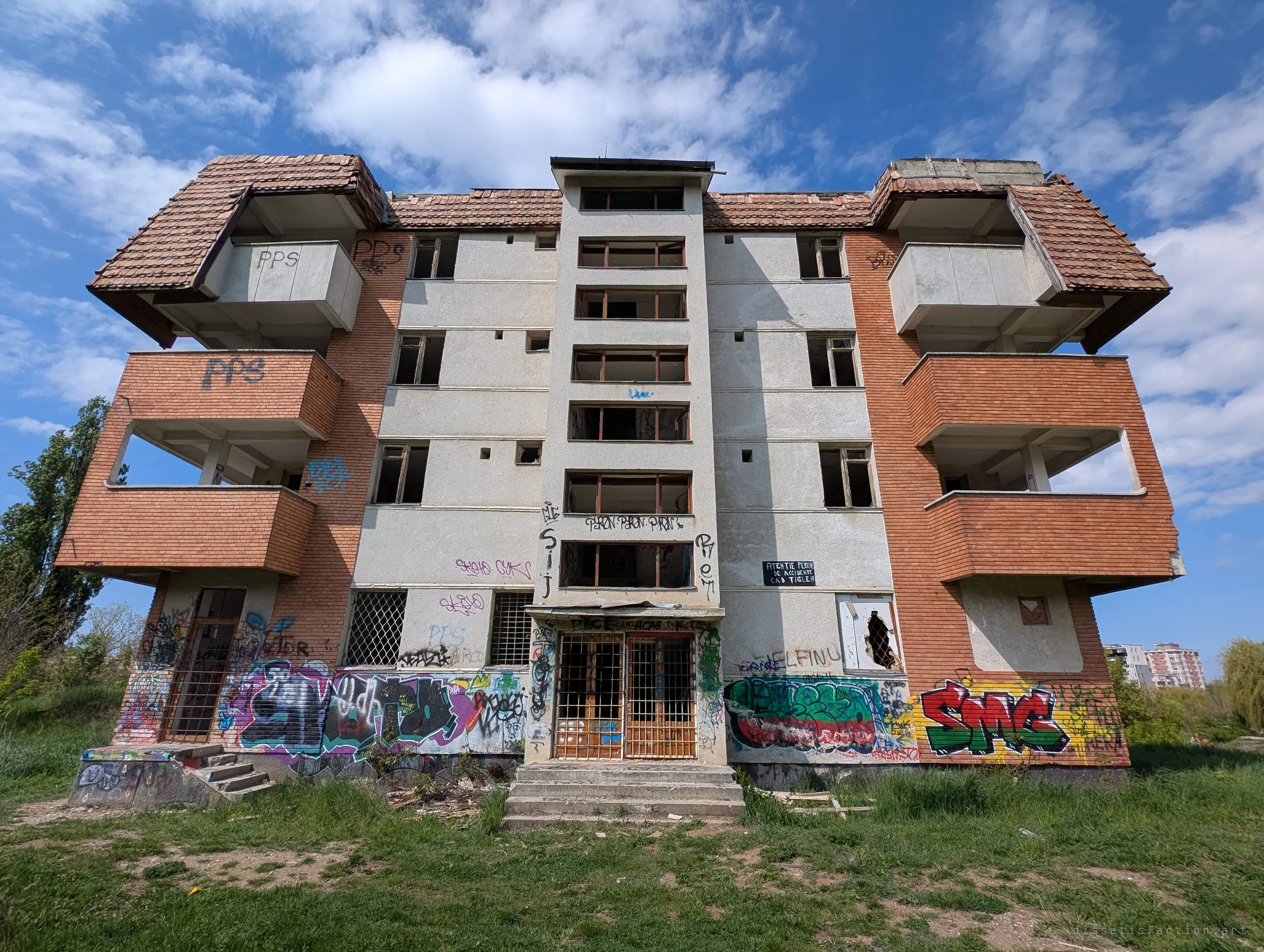 Wide front view of the abandoned block: a central stairwell tower of empty window frames flanked by brick balcony wings, the ground floor buried in graffiti. It shows the symmetrical layout of the whole building.