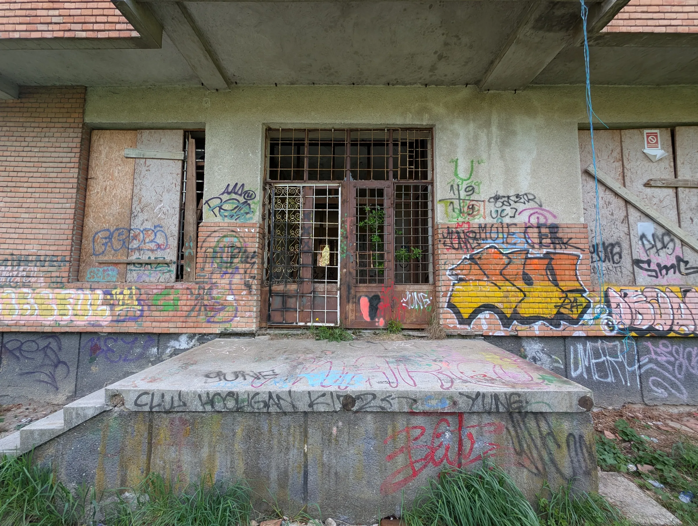 A barred entrance under a concrete overhang, with boarded windows on both sides and every surface tagged. A concrete loading step in front is layered with paint.