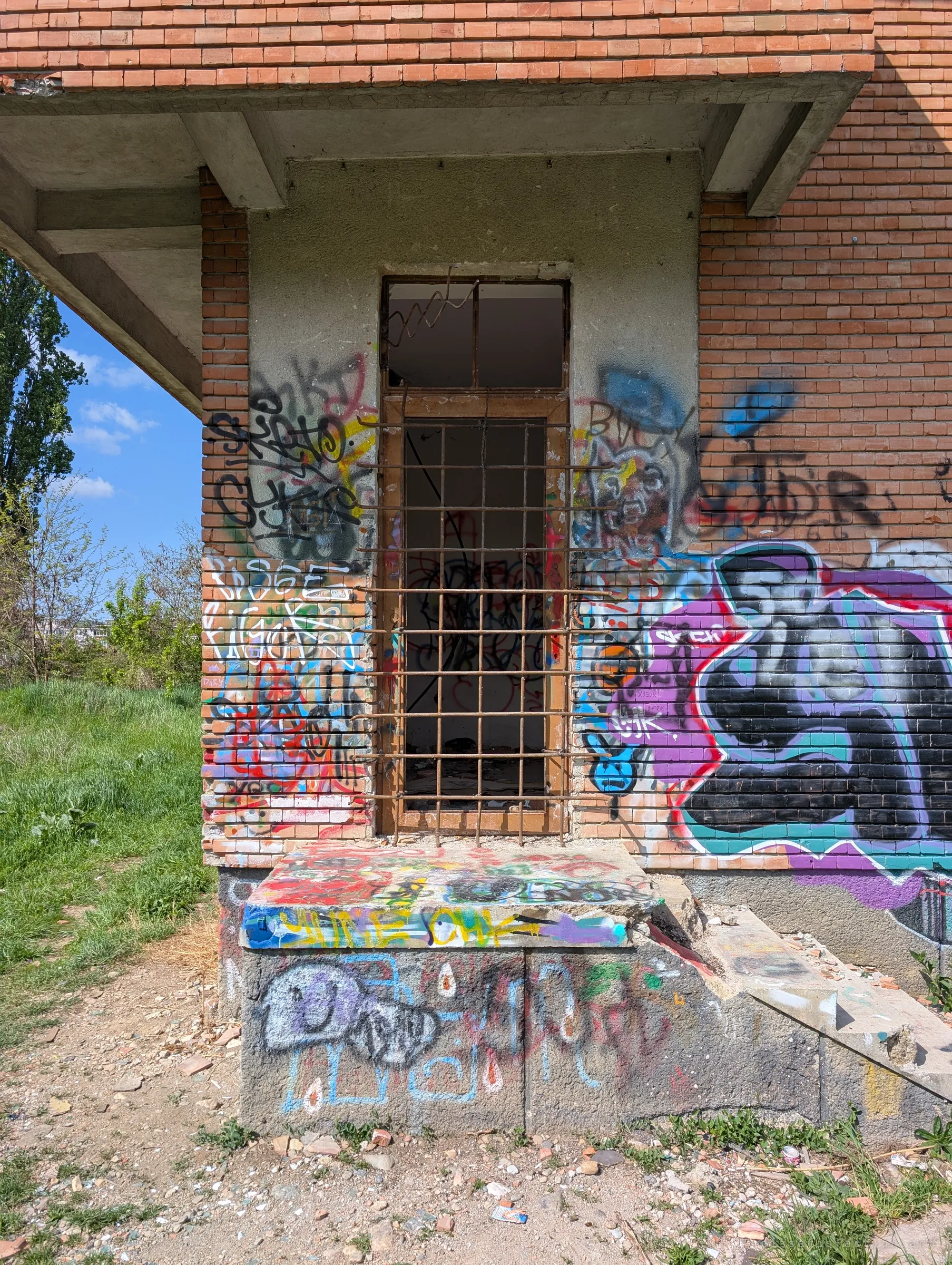 A tall side doorway with a bent iron grille, set in a brick wall thick with colourful graffiti. The raised step below hints at the way in.