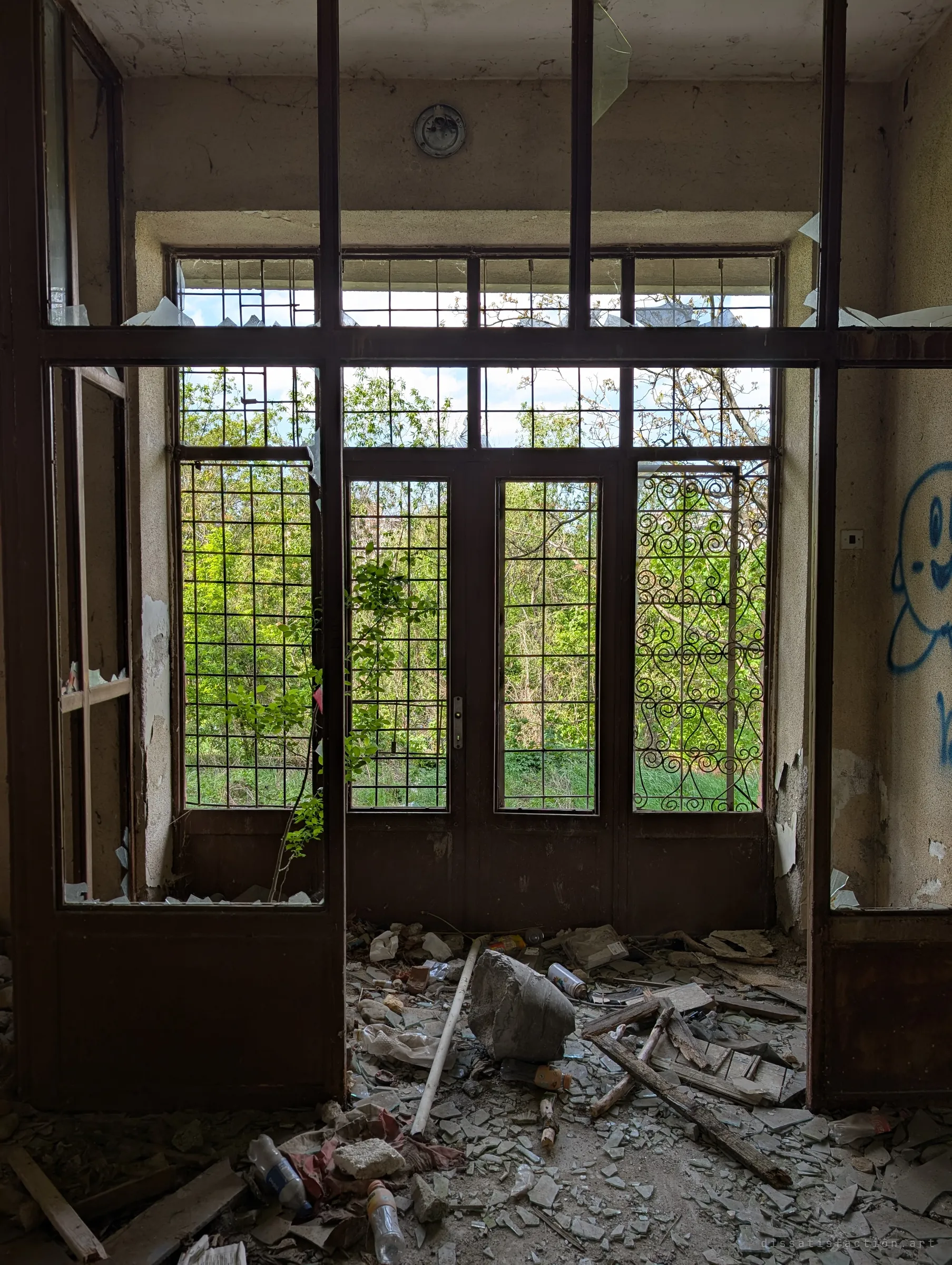 Looking out from inside through a gridded glass partition toward barred double doors, with green growth pressing against the glass. Shattered glass, rubble and bottles cover the floor.
