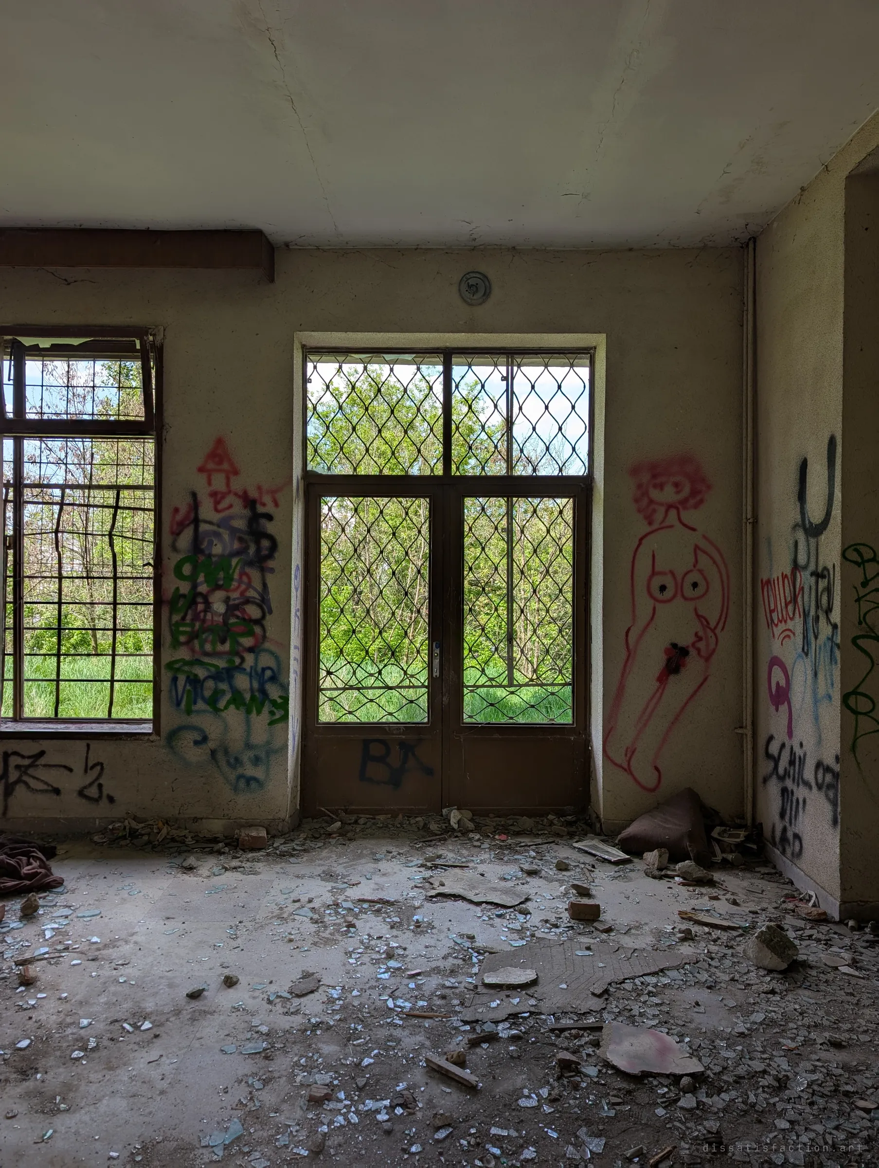 An empty ground-floor room with lattice-barred doors and a window opening onto trees. Crude graffiti figures cover the walls and broken glass litters the floor.