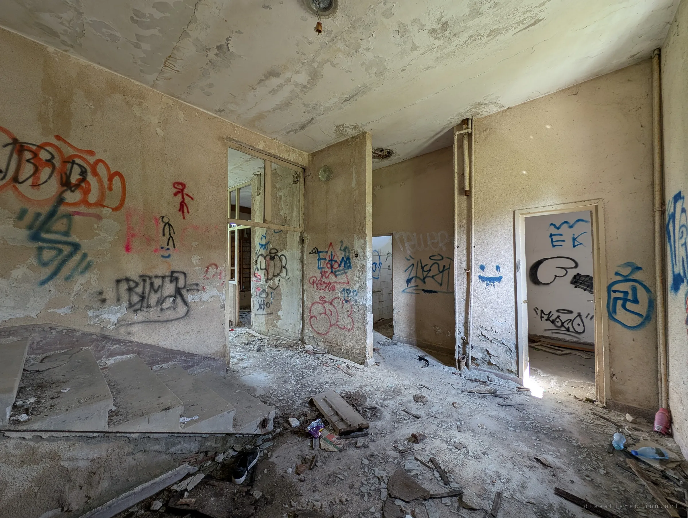 A wide interior hall with a curved concrete stair on the left and doorways leading off to tiled rooms. Peeling plaster and graffiti cover every wall.