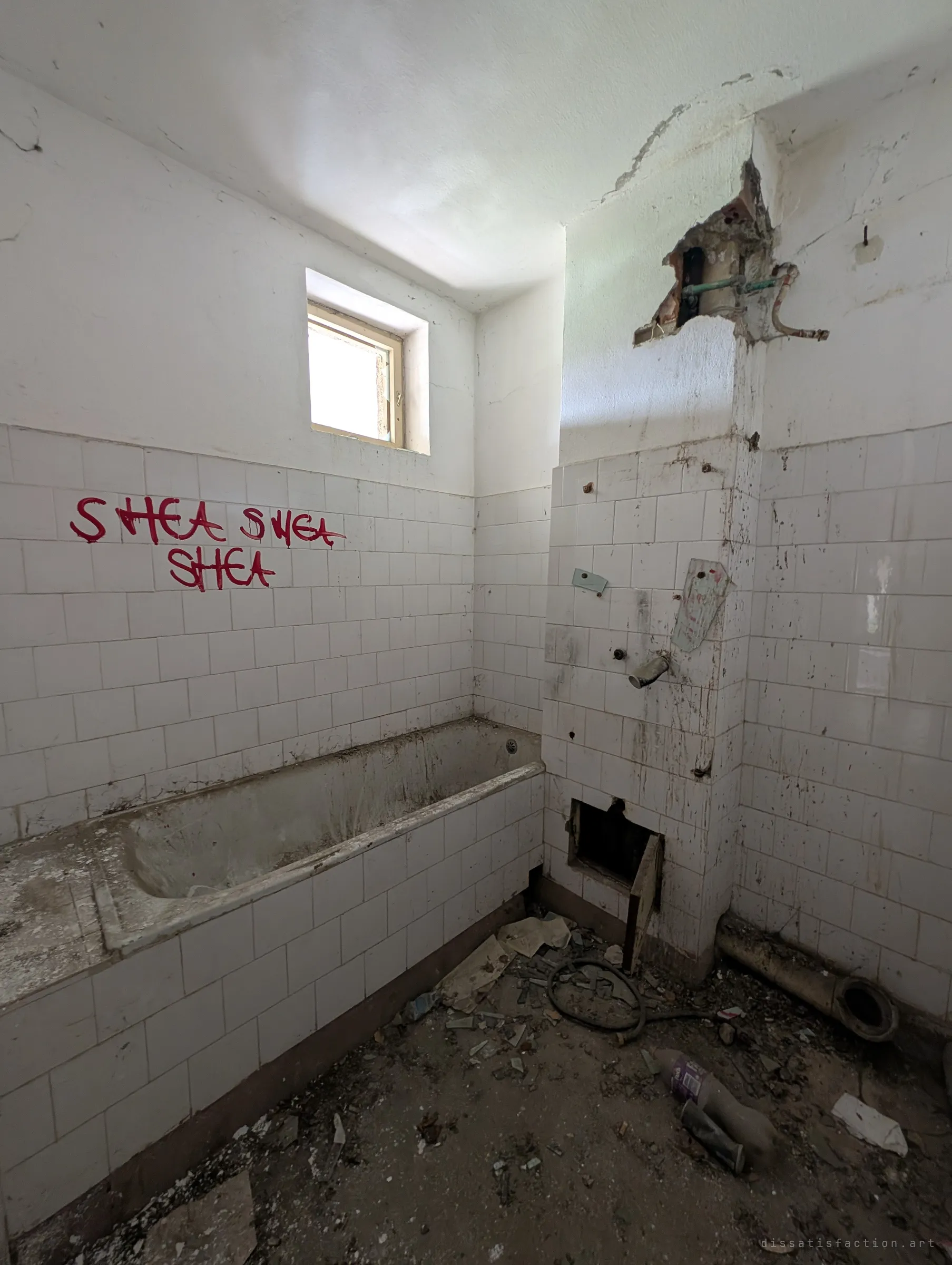 A stripped bathroom with a grimy tub, torn-out pipes and a hole hacked into the tiled wall. A small high window lets in flat daylight.