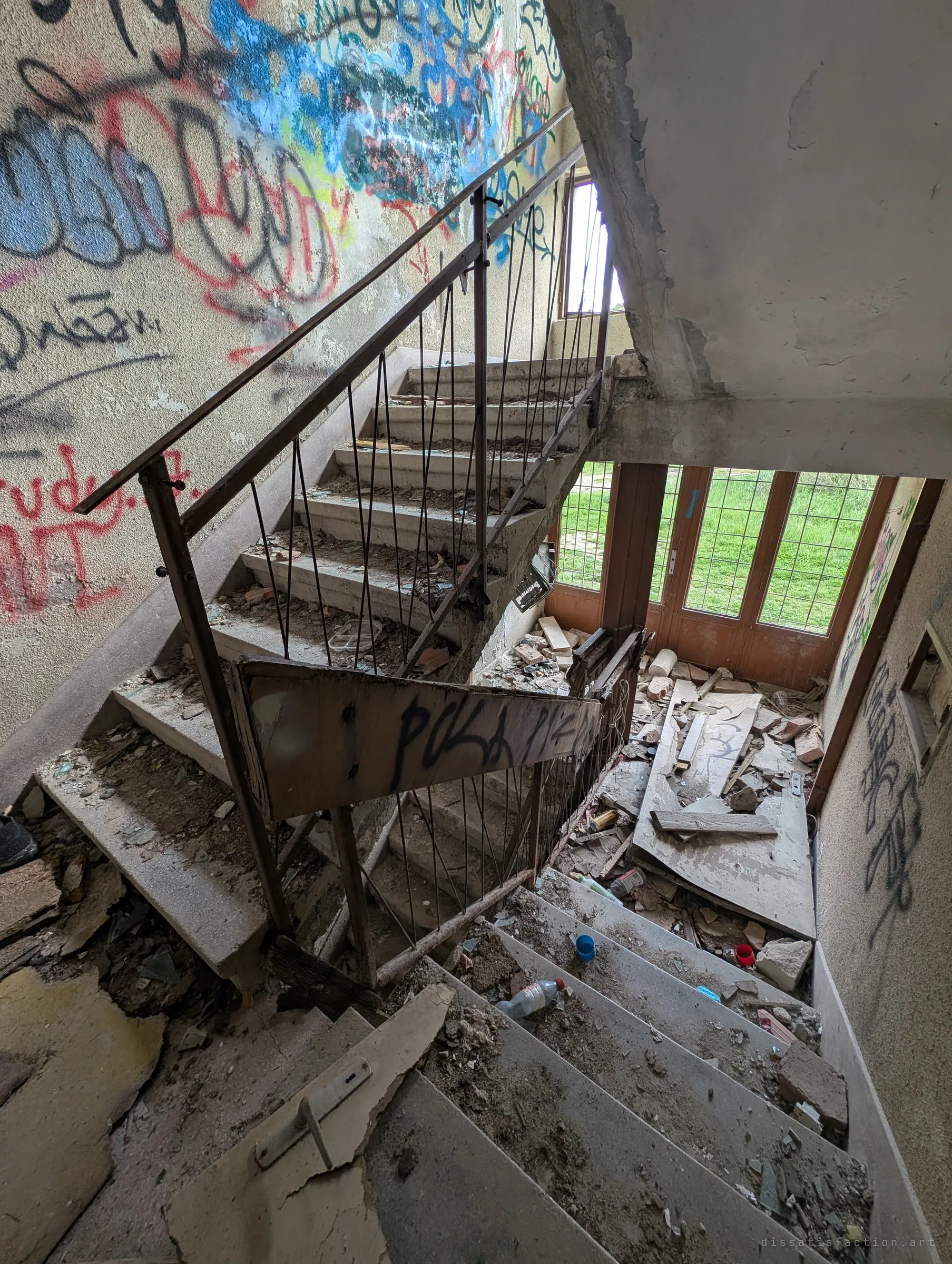 Looking down a stairwell with a thin metal railing, steps strewn with debris and glass, toward barred glass doors and grass outside. A ripped-out electrical box hangs from the wall.