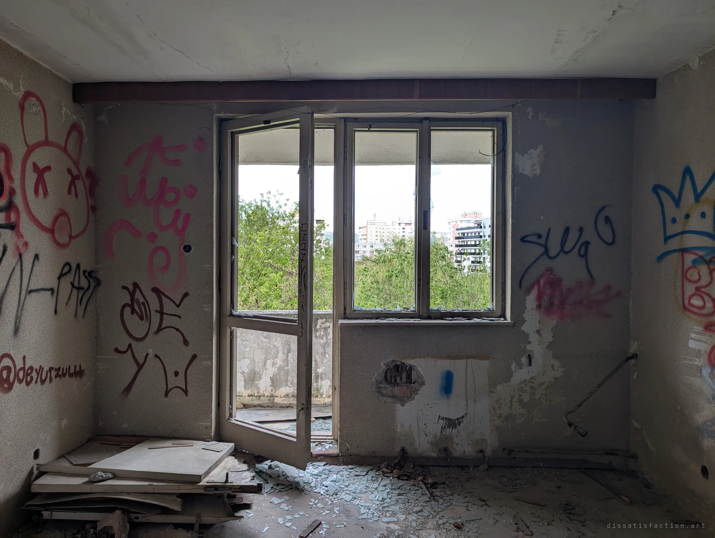 An upper-floor room with a balcony door hanging open and glass shards on the floor, walls covered in tags and cartoon faces. Beyond the window, trees and the city carry on as normal.