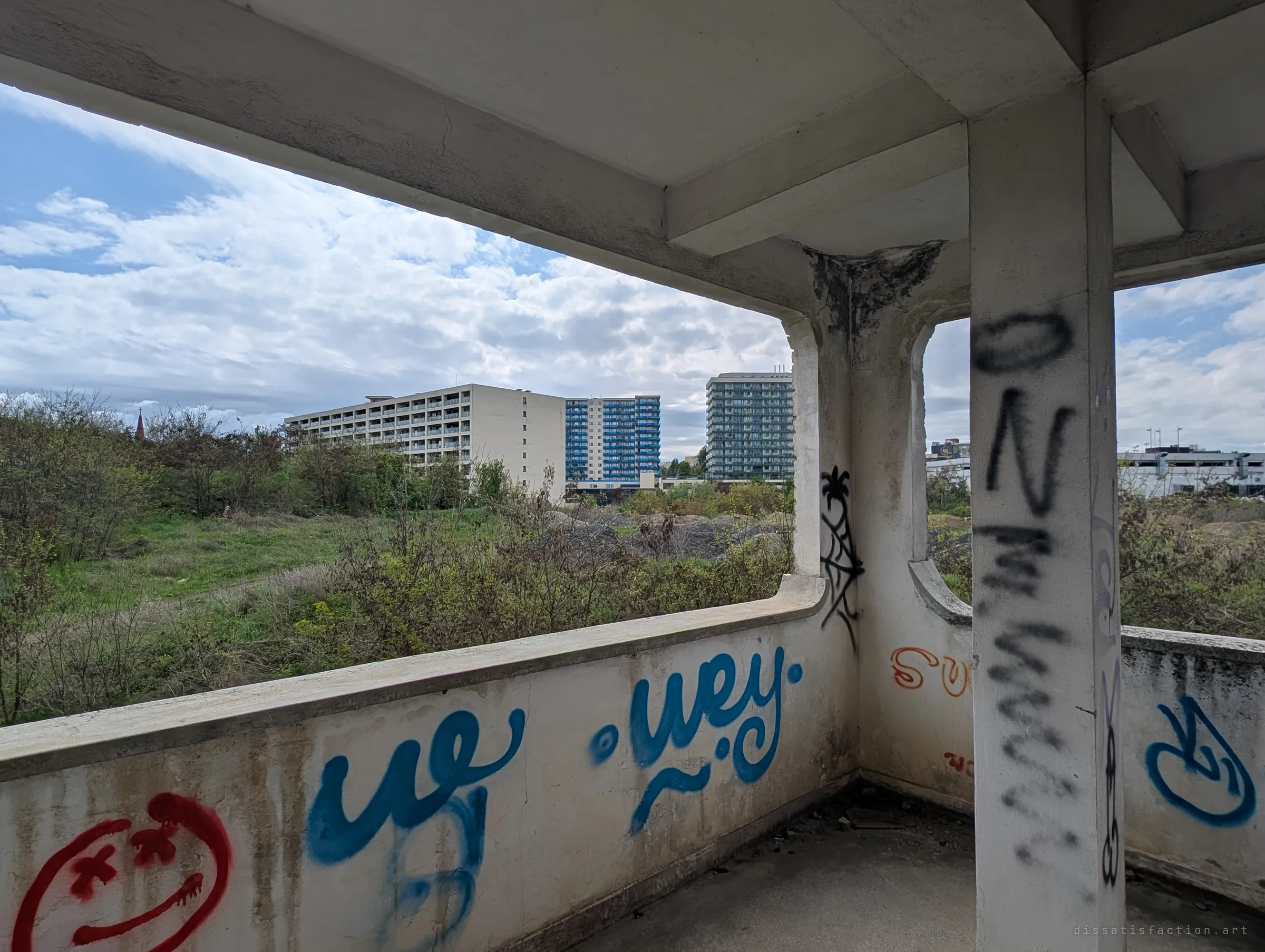 A graffiti-tagged concrete terrace with a thick column and low parapet, looking out over an overgrown lot. The open frame shows how exposed the upper levels of the building are.