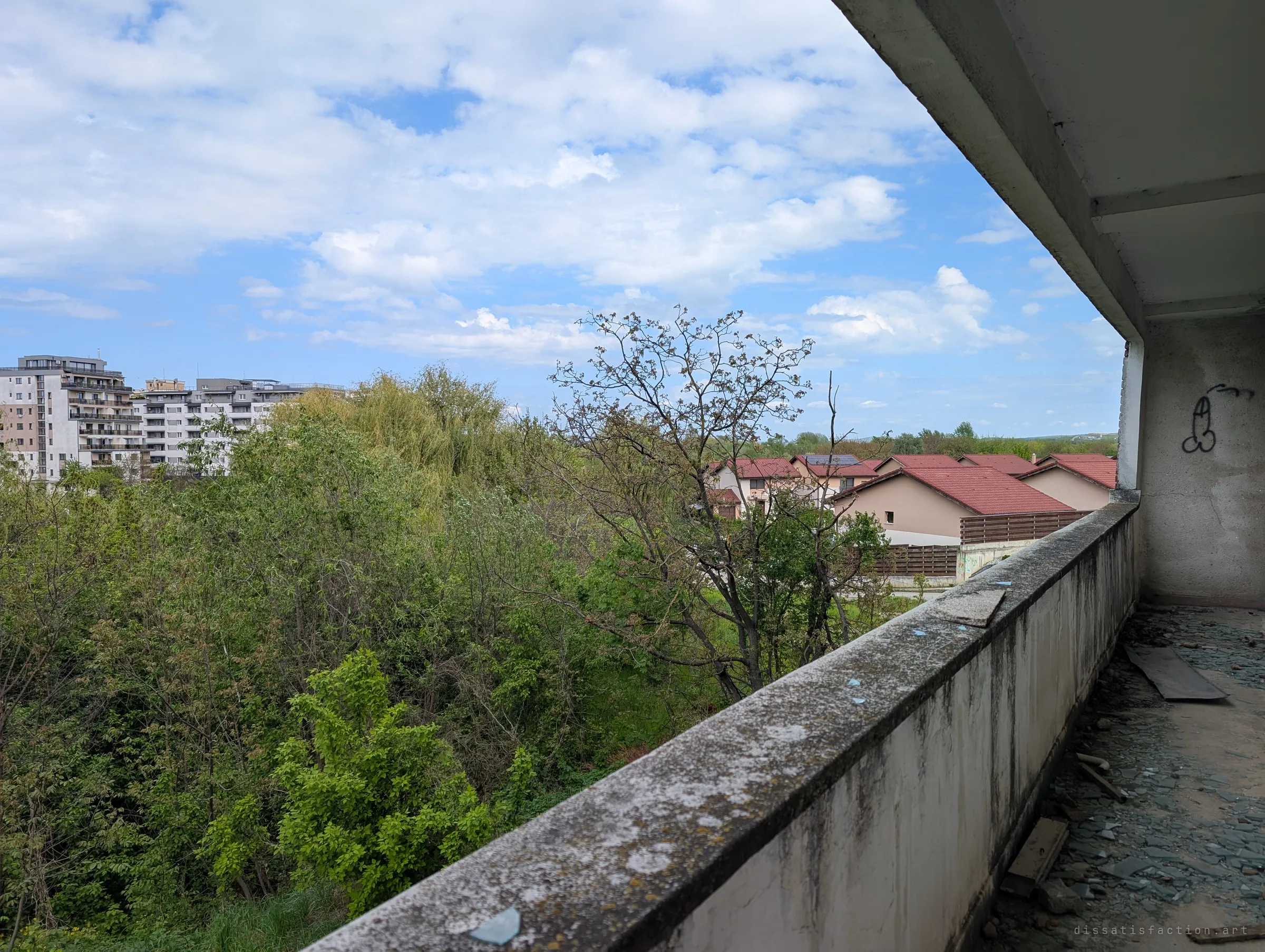 A long balcony strewn with broken glass runs along the building's edge, its parapet green with moss, above a dense stand of trees. It shows the height of the structure and how far the decay has spread.