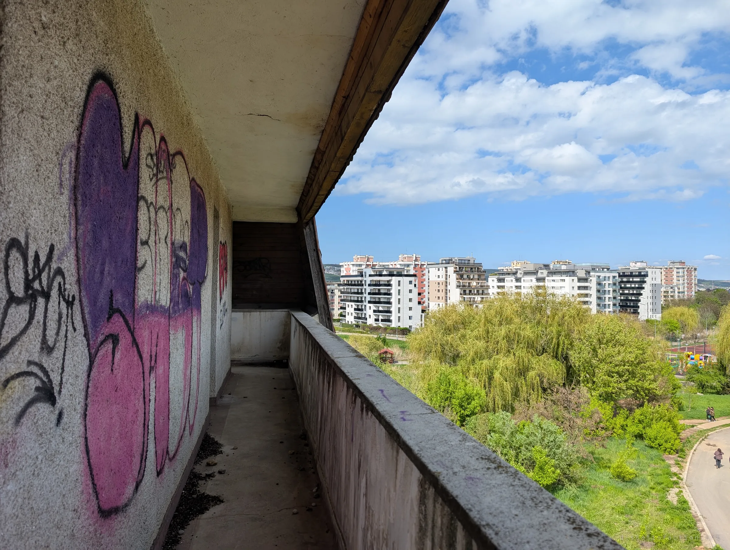 A narrow graffiti-covered balcony under a wooden eave, with purple and pink pieces along the wall and green parkland falling away beside it. The walkway shows the building's long, terraced upper floor.