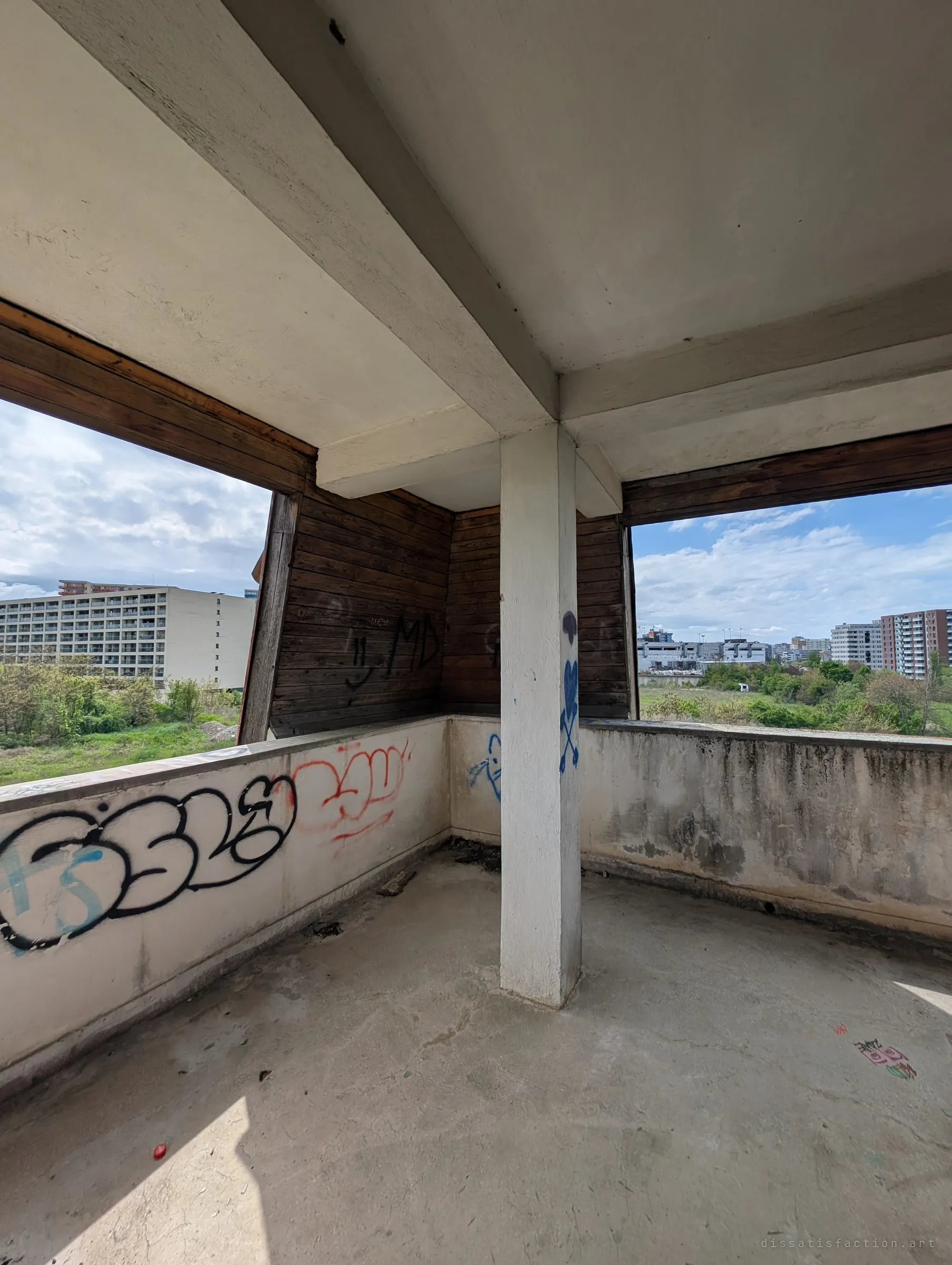 A corner room open to the sky on two sides, with a white concrete column, dark timber cladding and tagged low walls. It captures the building's stripped-back, half-outdoor upper storey.