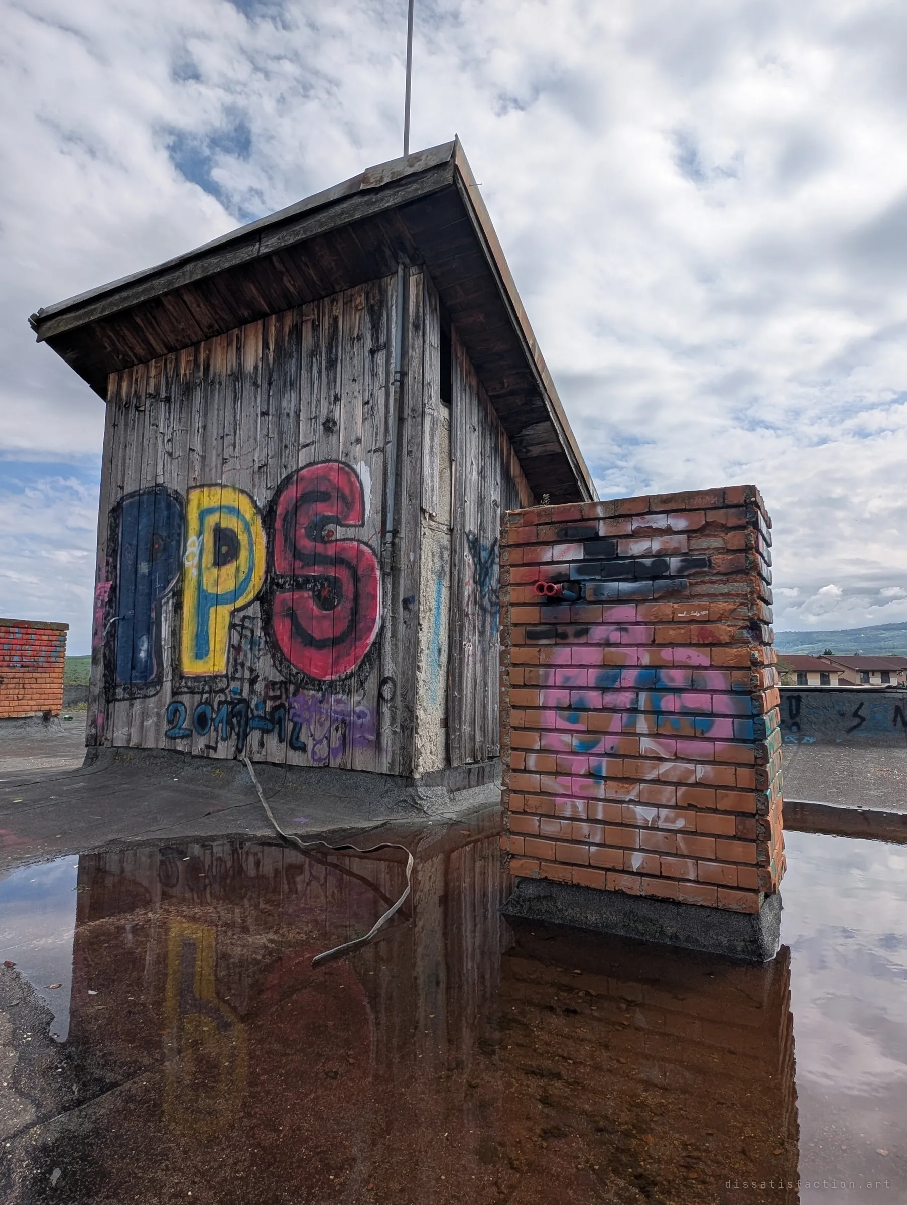 A weathered wooden rooftop hut with a big colourful letter piece and a brick chimney stack, both mirrored in a rust-red puddle on the flat roof. The reflection doubles the graffiti on the abandoned roof.