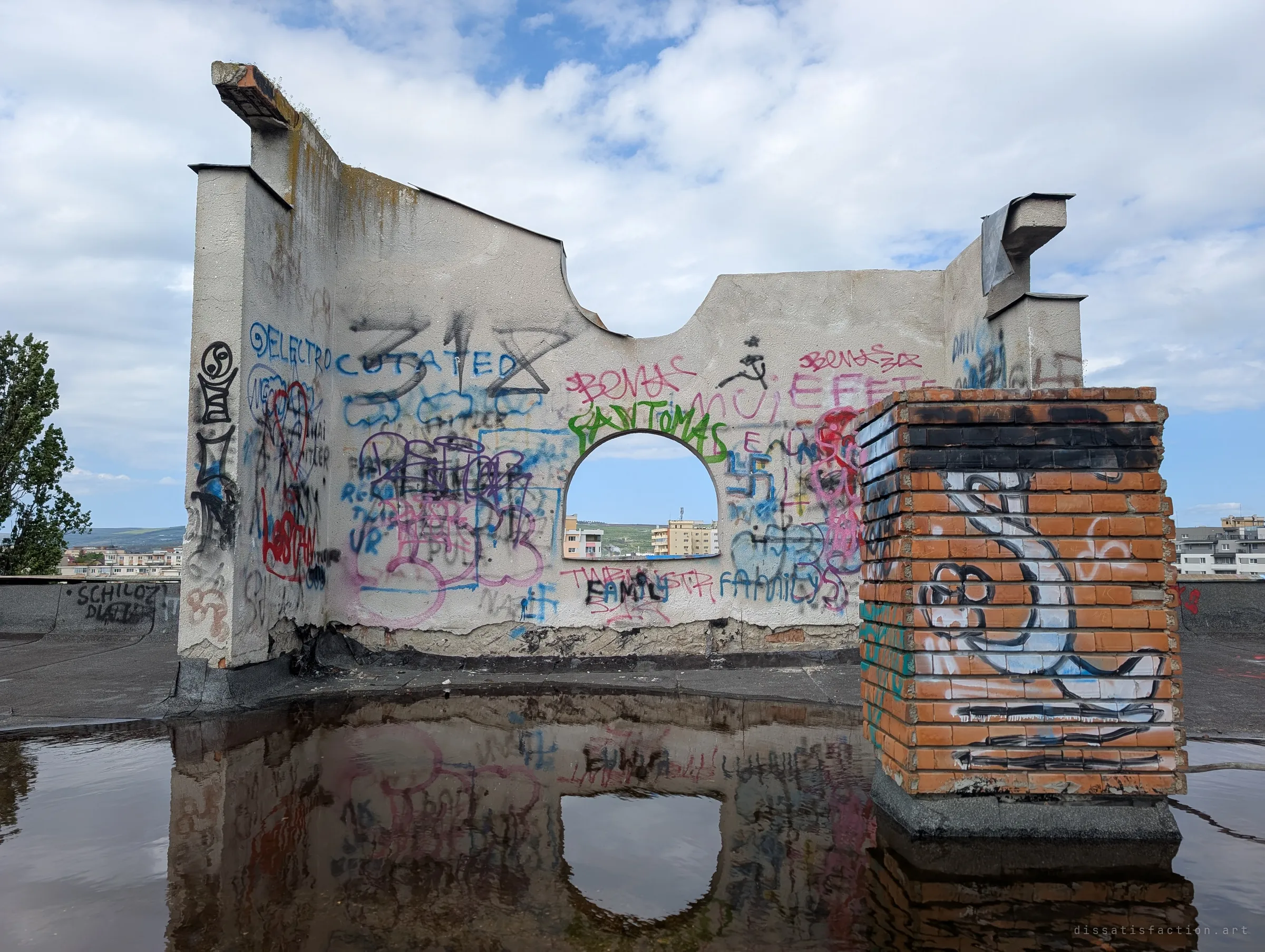 A freestanding rooftop wall with an arched opening, covered in layers of tags, reflected in a large puddle beside a painted brick chimney. It shows how heavily visited and written-over the roof has become.