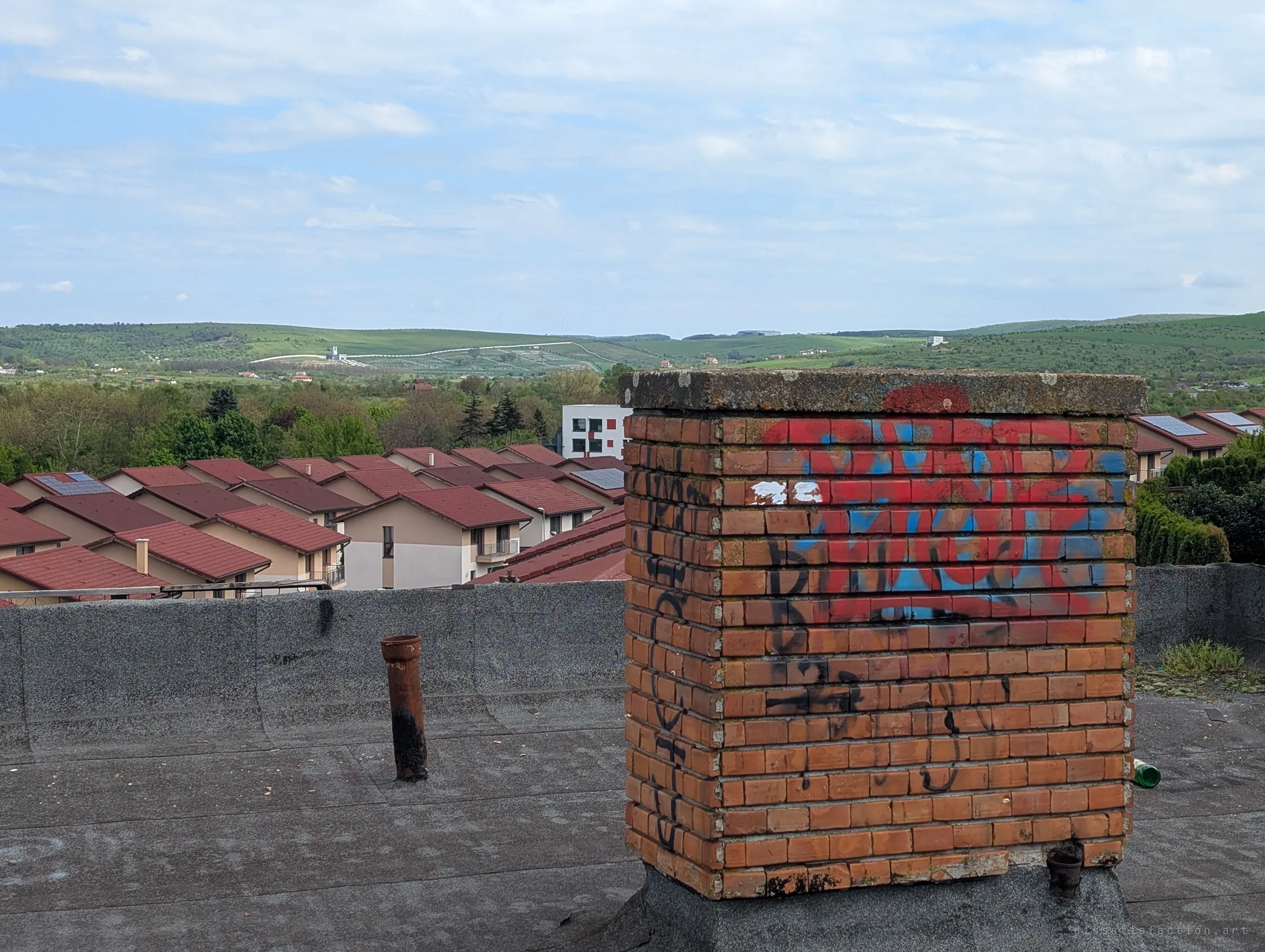 A graffiti-tagged brick chimney and a rusty vent pipe on the tar roof, with rows of red-roofed houses and green hills behind. The rooftop details sit against the quiet suburb around the ruin.