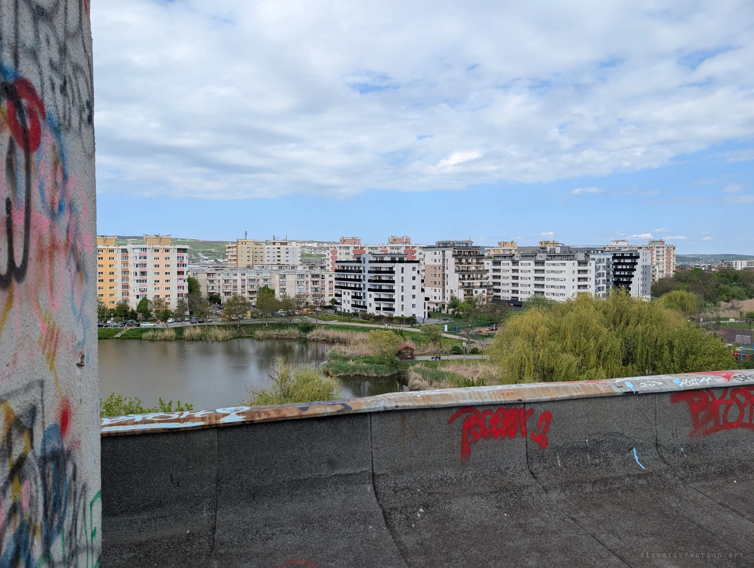 A tagged roof parapet and graffiti-covered pillar frame a view over a pond ringed by reeds and apartment blocks. It shows how close the ruin sits to a busy living neighbourhood.