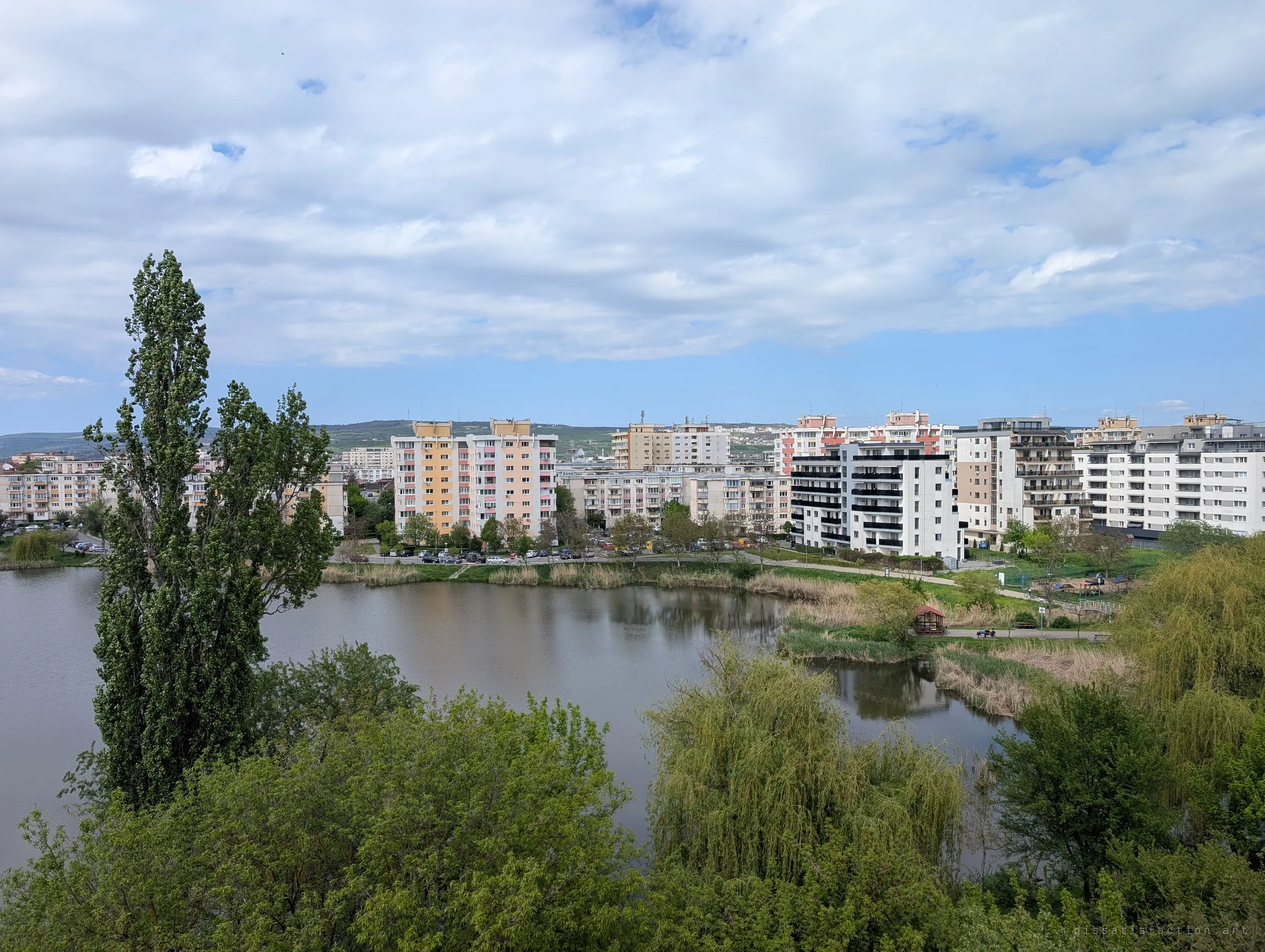 A tall poplar and willows in front of a still pond, with rows of apartment blocks and hills beyond under a big cloudy sky. The view from the top floor looks out over the surrounding district.