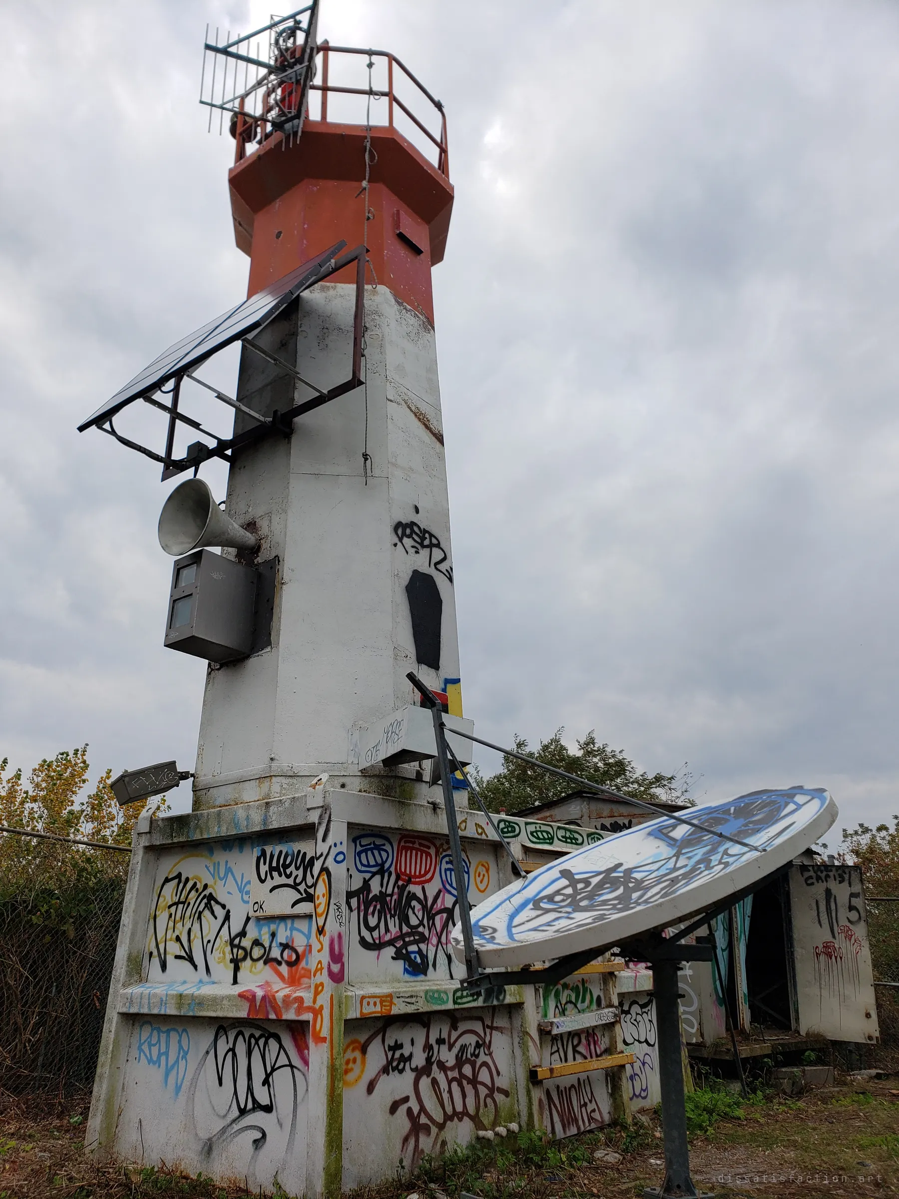 A graffiti-covered white light tower with a red lantern top, solar panel and foghorn, shot from below under grey cloud. A tagged satellite dish leans in the foreground.