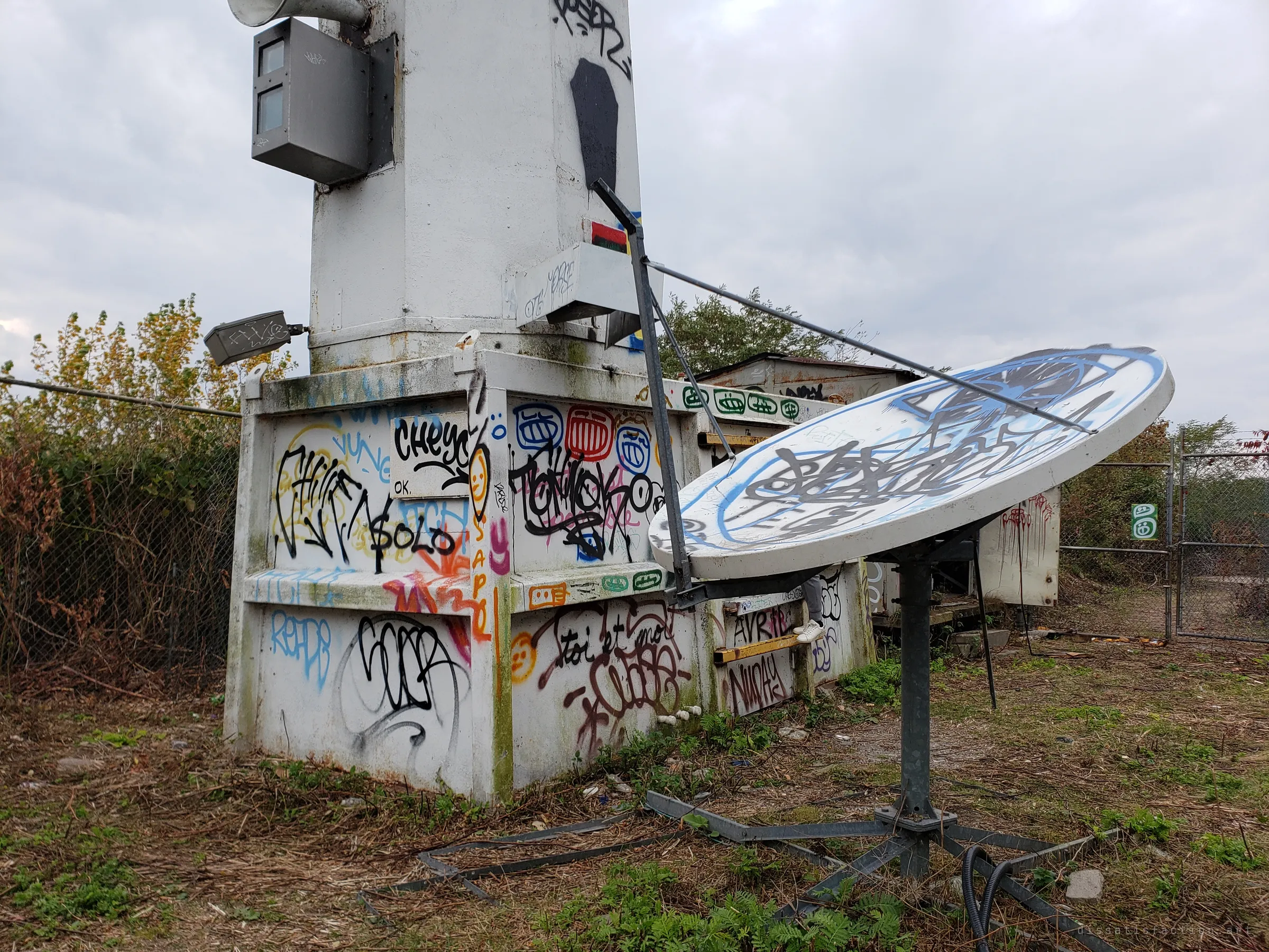 The tagged concrete base of the light tower beside a graffiti-covered satellite dish, inside a chain-link fence overgrown with brush.