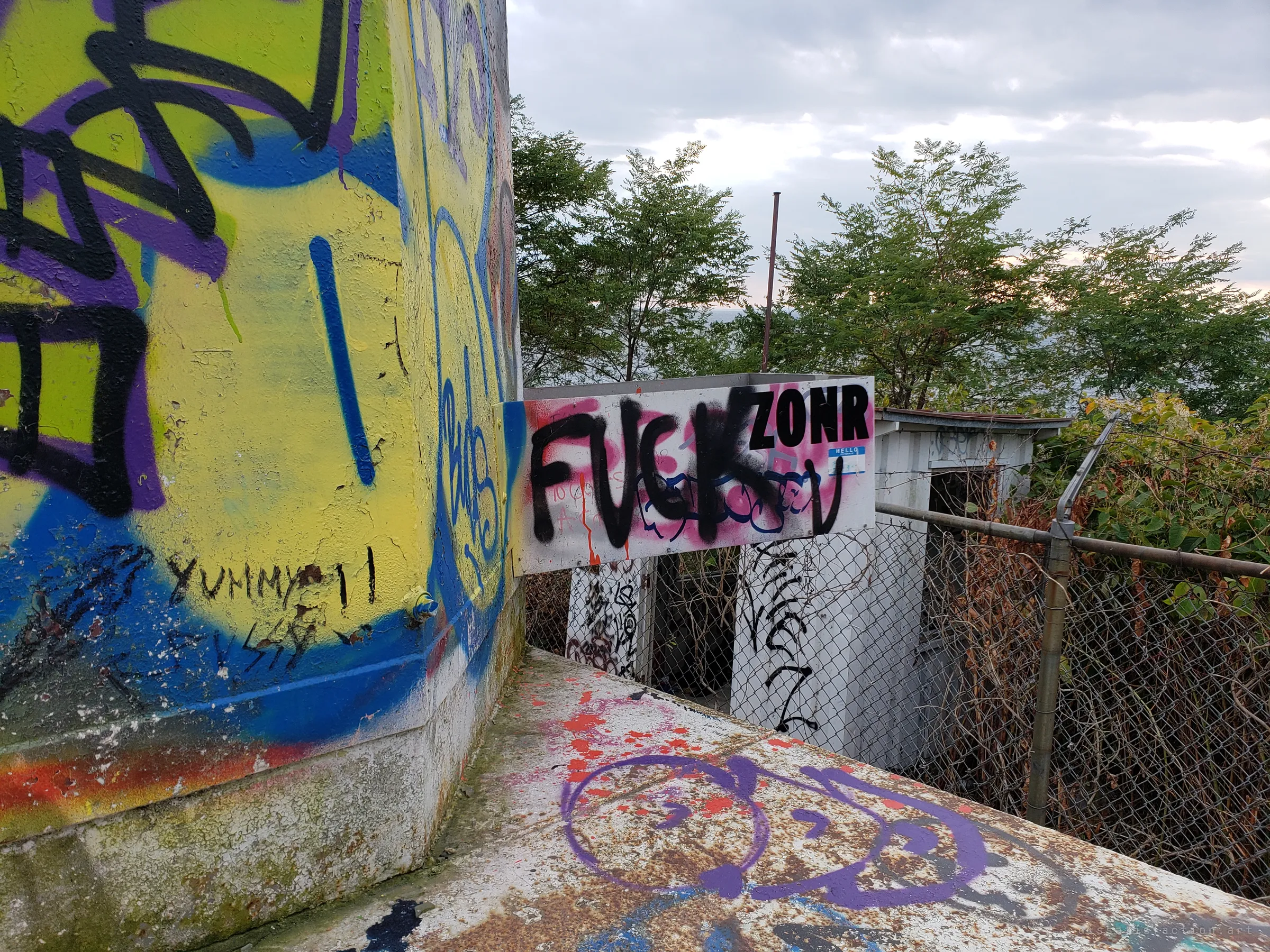 The painted tower wall meets a rusted ledge, with a sprayed-over sign and a small outbuilding behind a chain-link fence and trees.