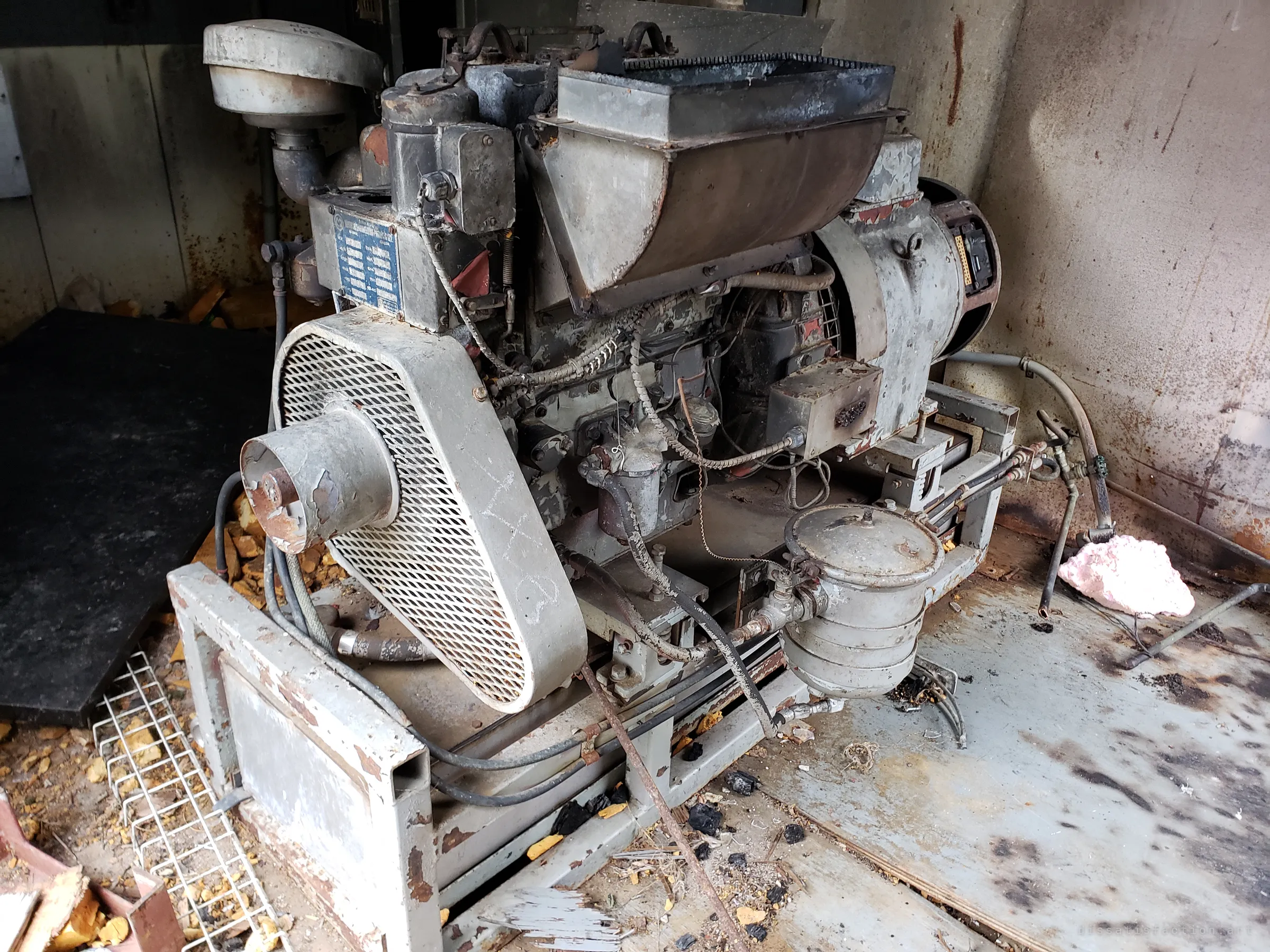 A soot-streaked backup generator engine sits abandoned on the rusted shed floor among burnt debris and scraps of insulation.