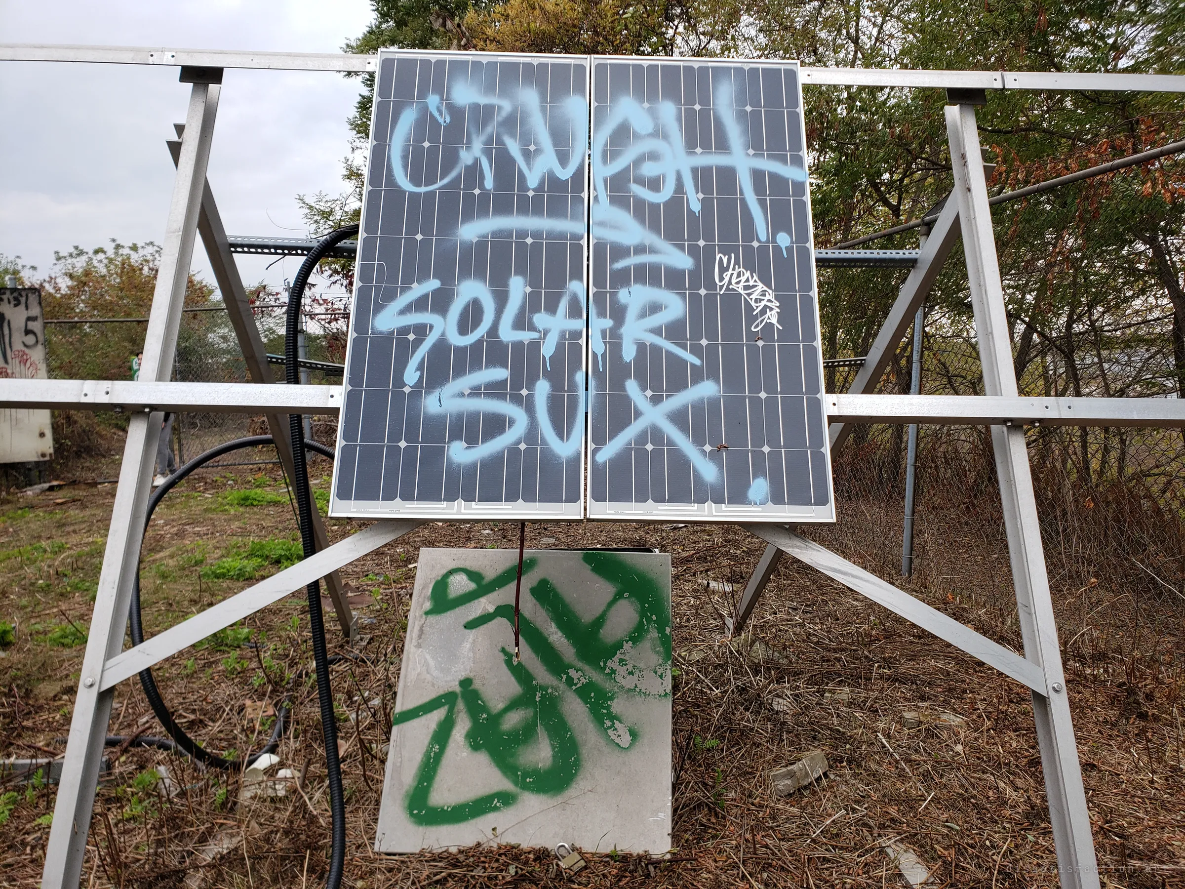A pair of solar panels on a steel frame, sprayed with a sarcastic tag, above a padlocked grey box marked in green paint.