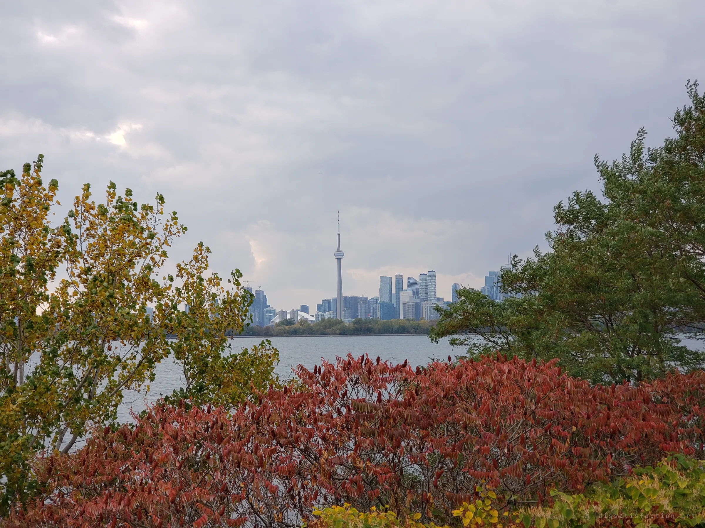A city skyline rises across grey water, framed by autumn foliage in red and yellow under heavy cloud.