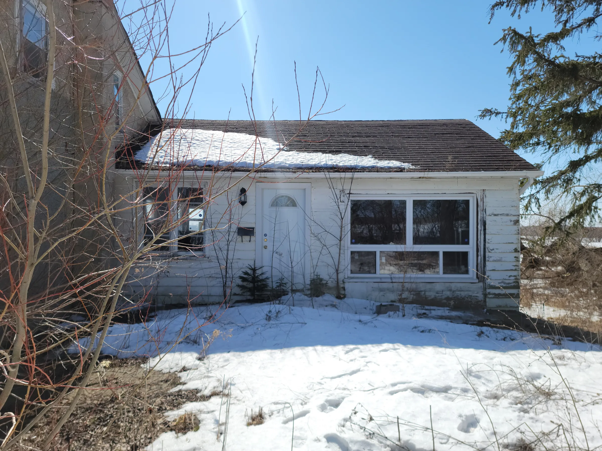 Snow-covered front yard of a white clapboard side wing with a picture window and a small front door, bare saplings crowding the frame. It sets the scene of a quiet rural house left to the weather.