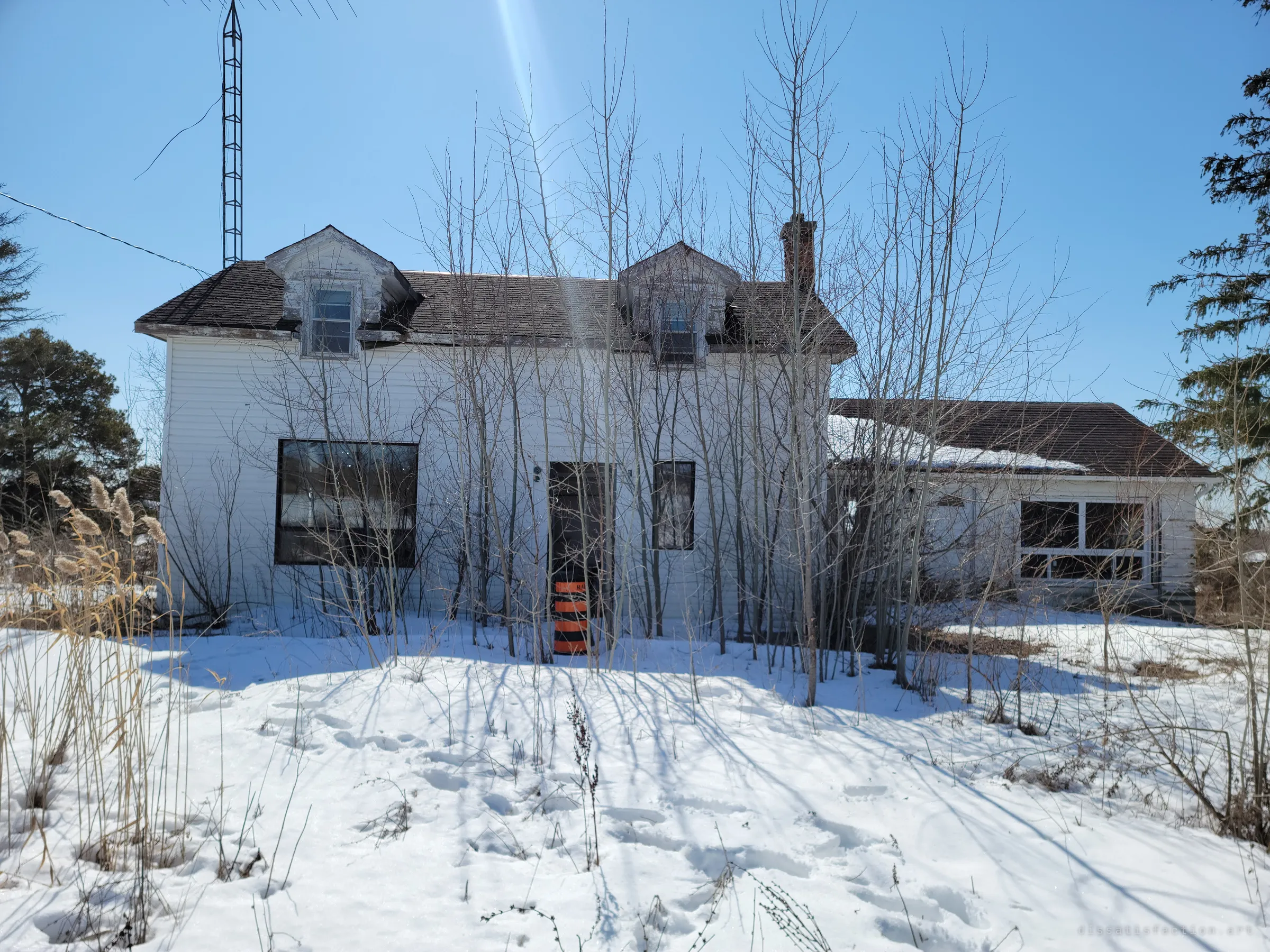A white two-storey farmhouse with twin dormers stands behind a thicket of bare saplings, an orange road barrel parked at its front door. The deep snow and overgrowth show how long it has sat empty.