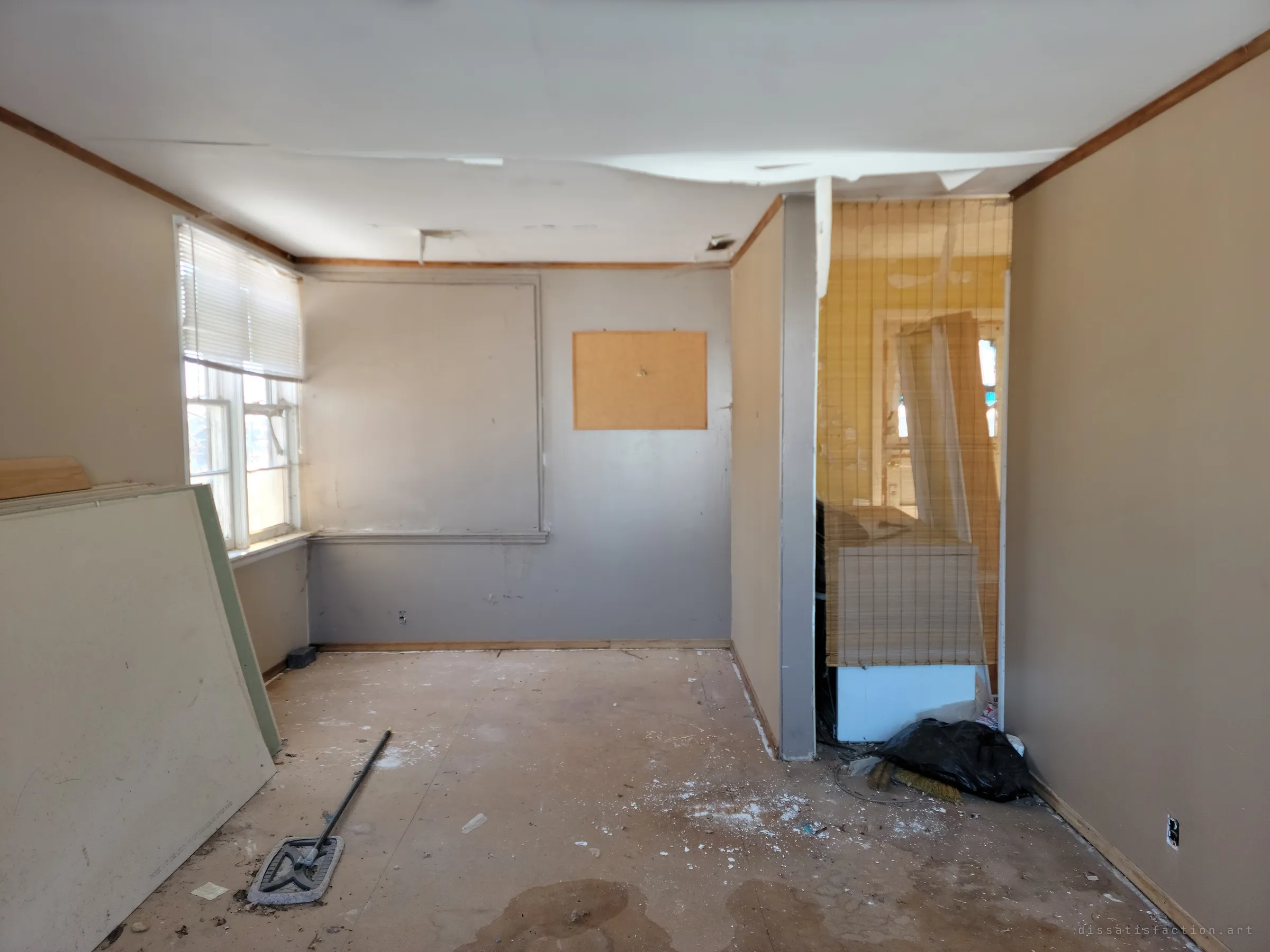 An emptied room with sagging ceiling paper, a bare corkboard on the wall and a mop left on the stained subfloor. A bamboo blind hangs across the doorway into the yellow room beyond.