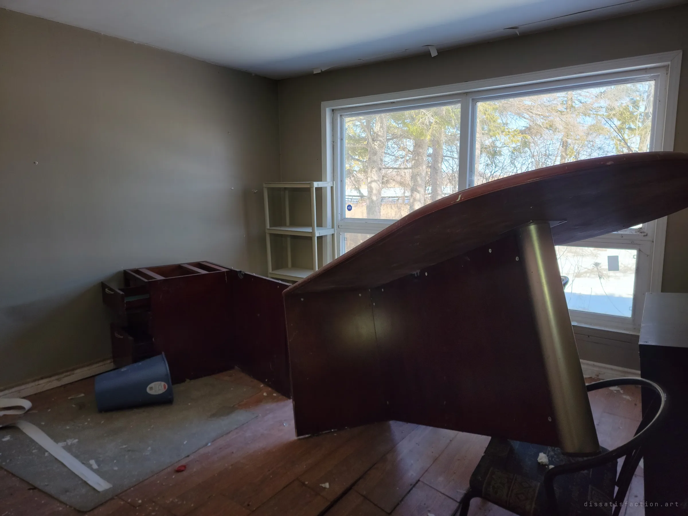 A dim room where a large wooden table lies tipped on its side before a bright picture window. Dismantled office furniture and a toppled bin suggest someone started clearing out and never finished.