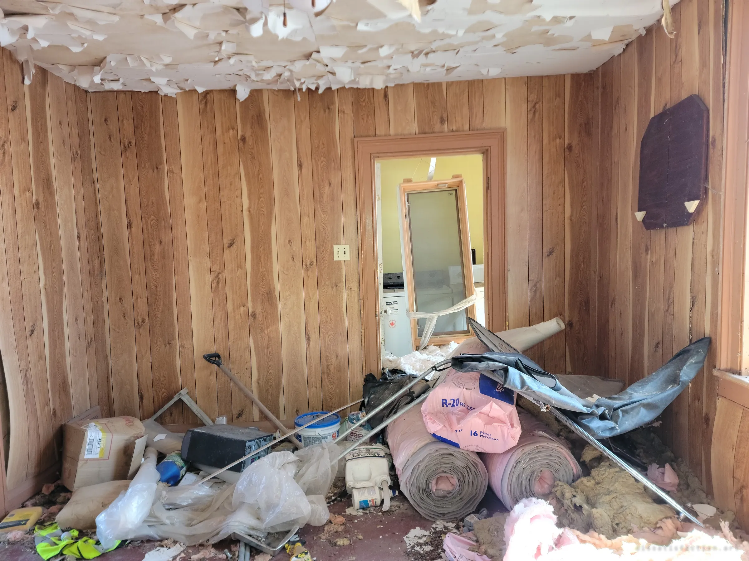 A wood-panelled room piled with insulation rolls, paint pails and renovation leftovers under a peeling ceiling. Through the doorway a washer and dryer stand in a yellow laundry room.