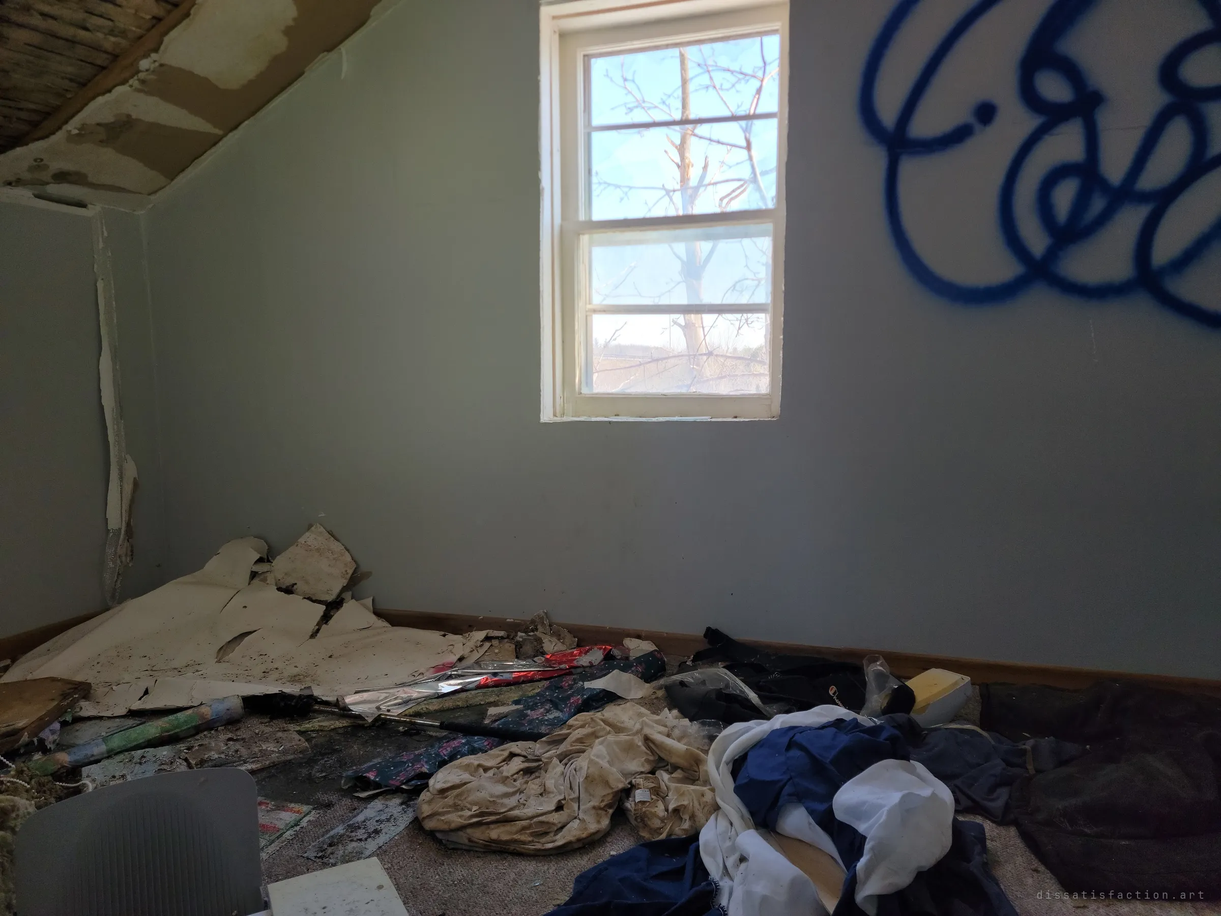 An upstairs room under a sloped ceiling, blue spray-paint tag on the wall and clothes and fallen plaster heaped on the carpet. The light tag is the only sign of visitors.