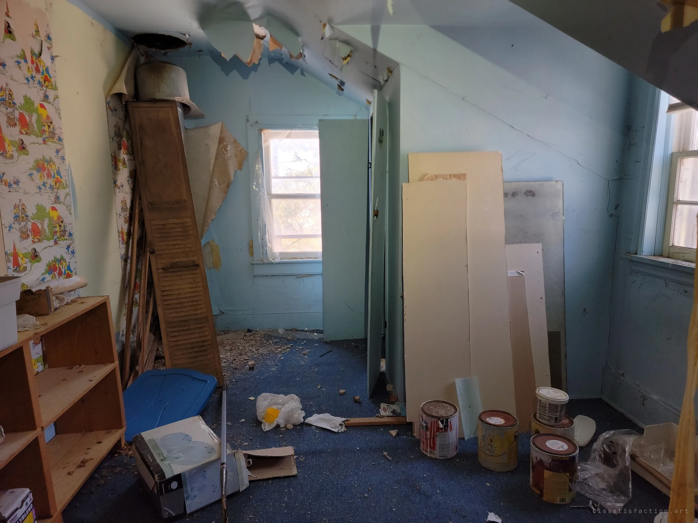 An attic bedroom with cartoon-mushroom wallpaper, blue carpet, leaning boards and open paint cans. The childhood wallpaper makes the abandonment feel personal.