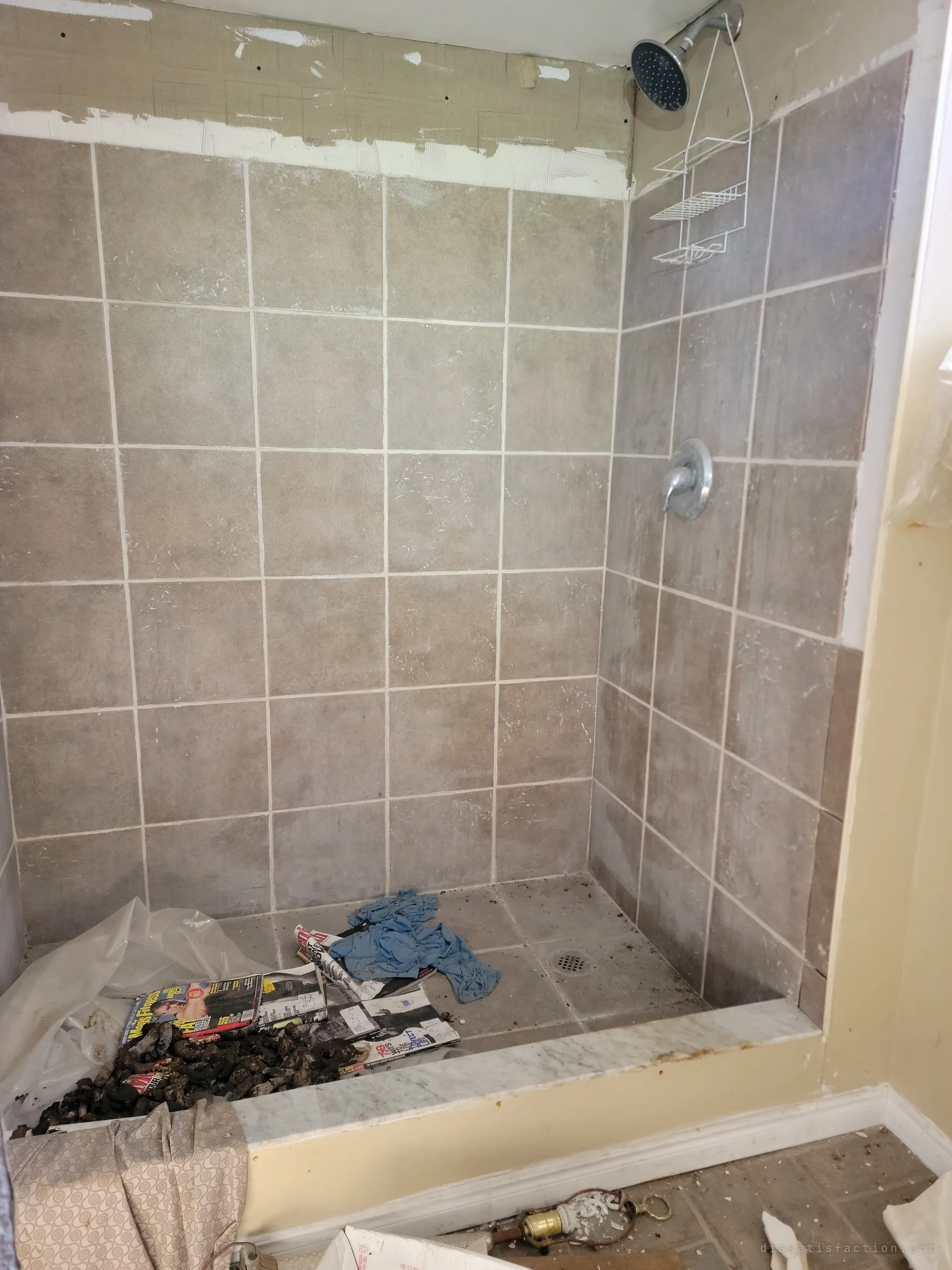 A half-renovated tiled shower stall with old magazines, a rag and debris dumped in its base. Renovation work stopped mid-job.