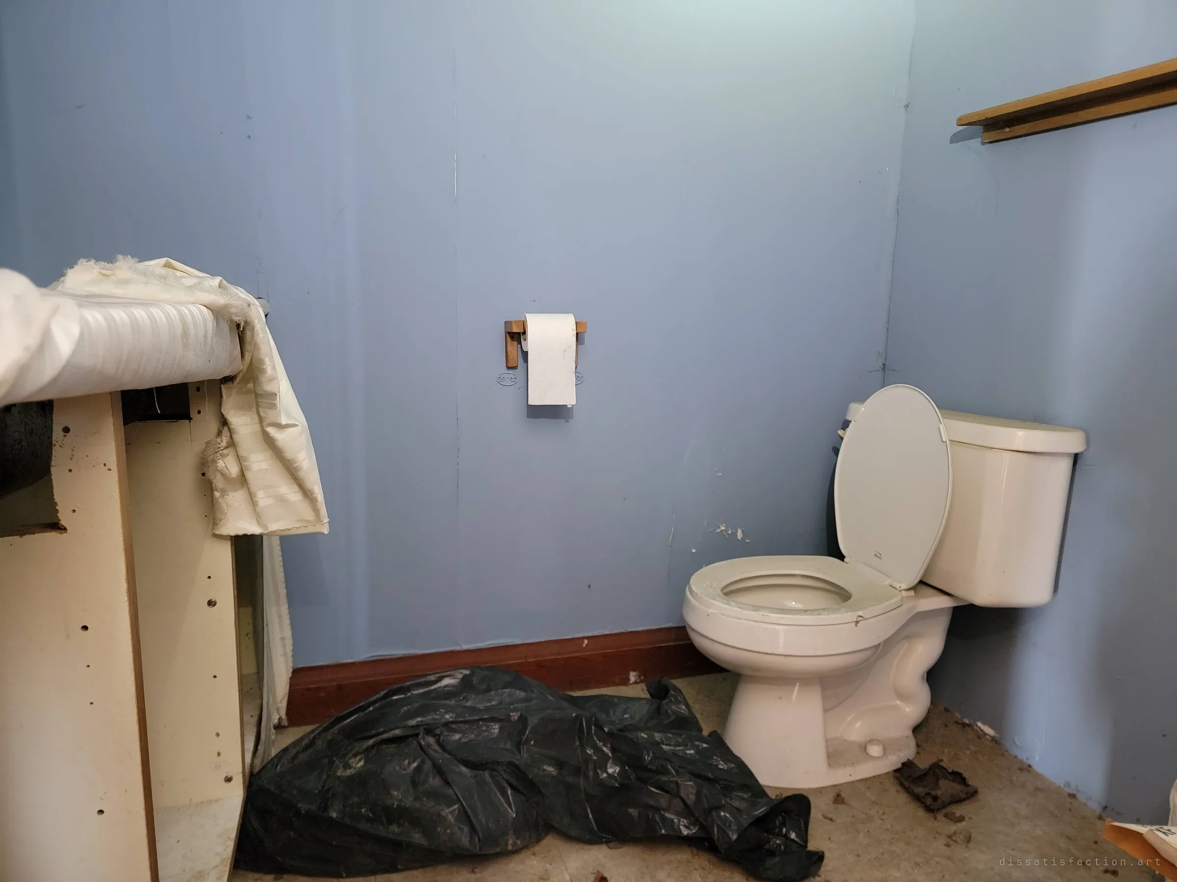 A small blue bathroom with a toilet, a lone roll of paper still on its holder and a torn black bag on the floor. The paper roll left in place makes the abandonment feel sudden.