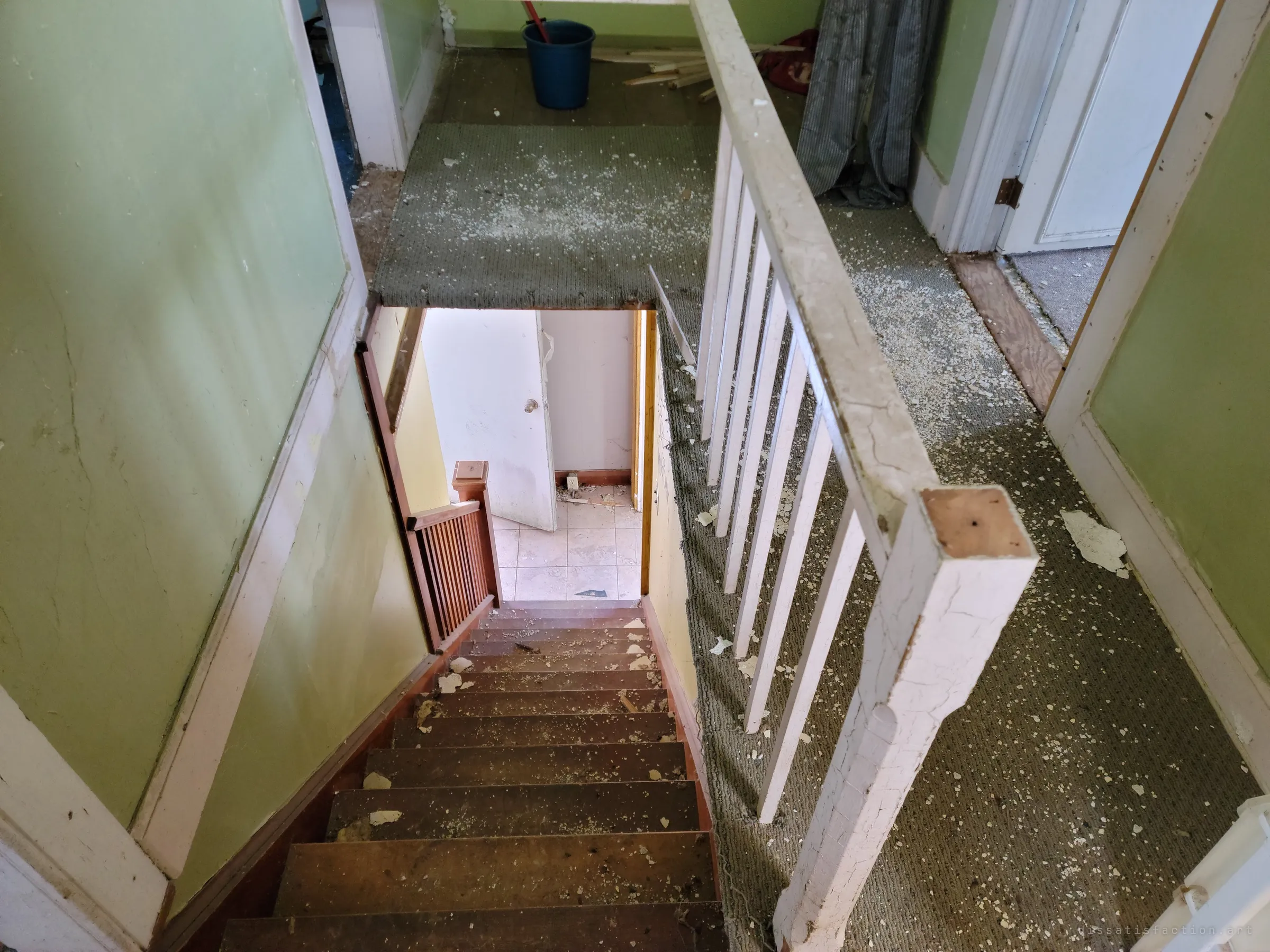 Looking down a steep wooden staircase from an upstairs landing littered with fallen plaster, past a cracked white banister. A door stands open at the bottom onto a tiled room.