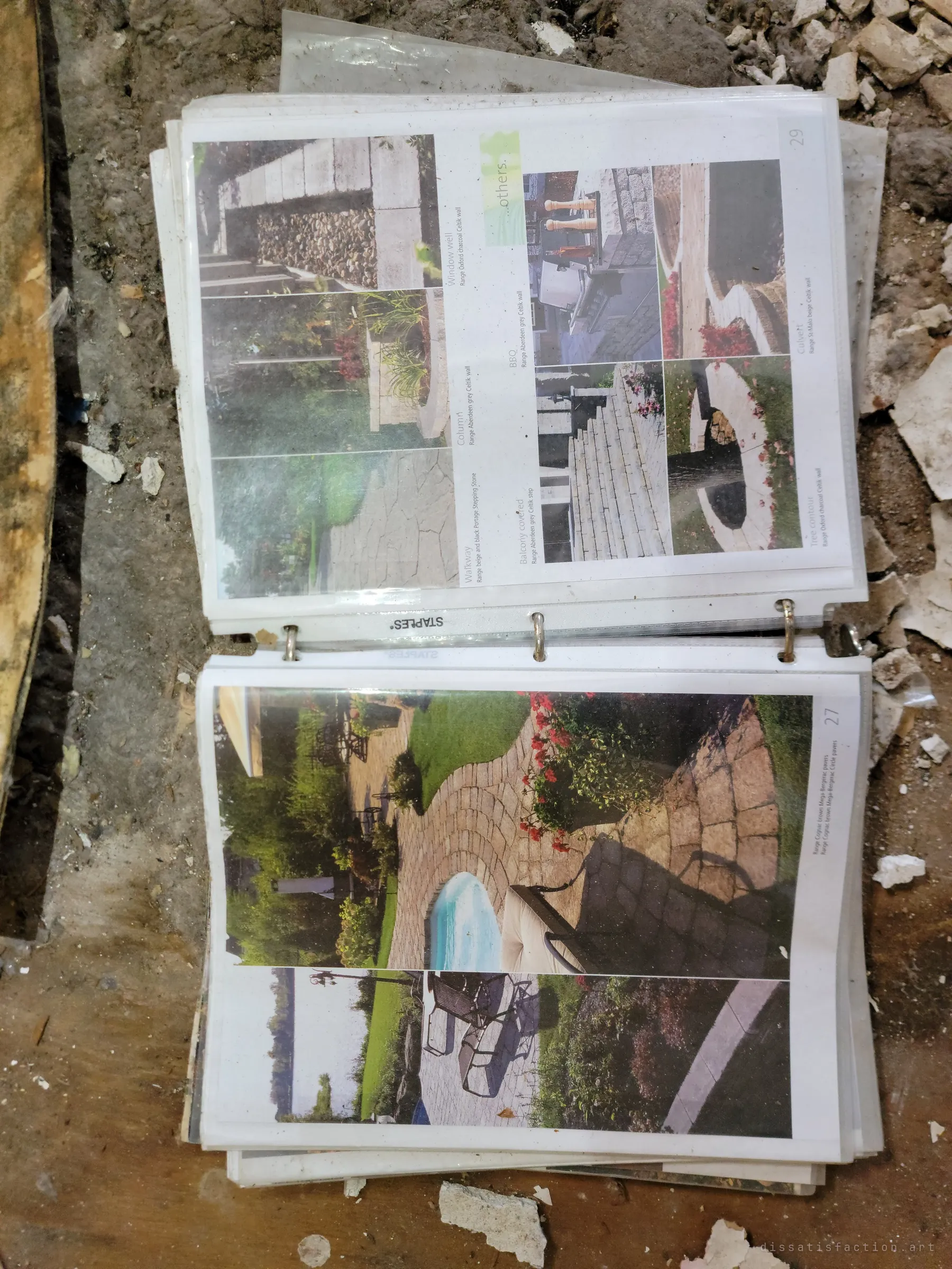 A binder of landscaping product brochures lying open in the rubble, showing patios, walls and garden paths. Glossy plans for a tidy yard, abandoned in the debris.