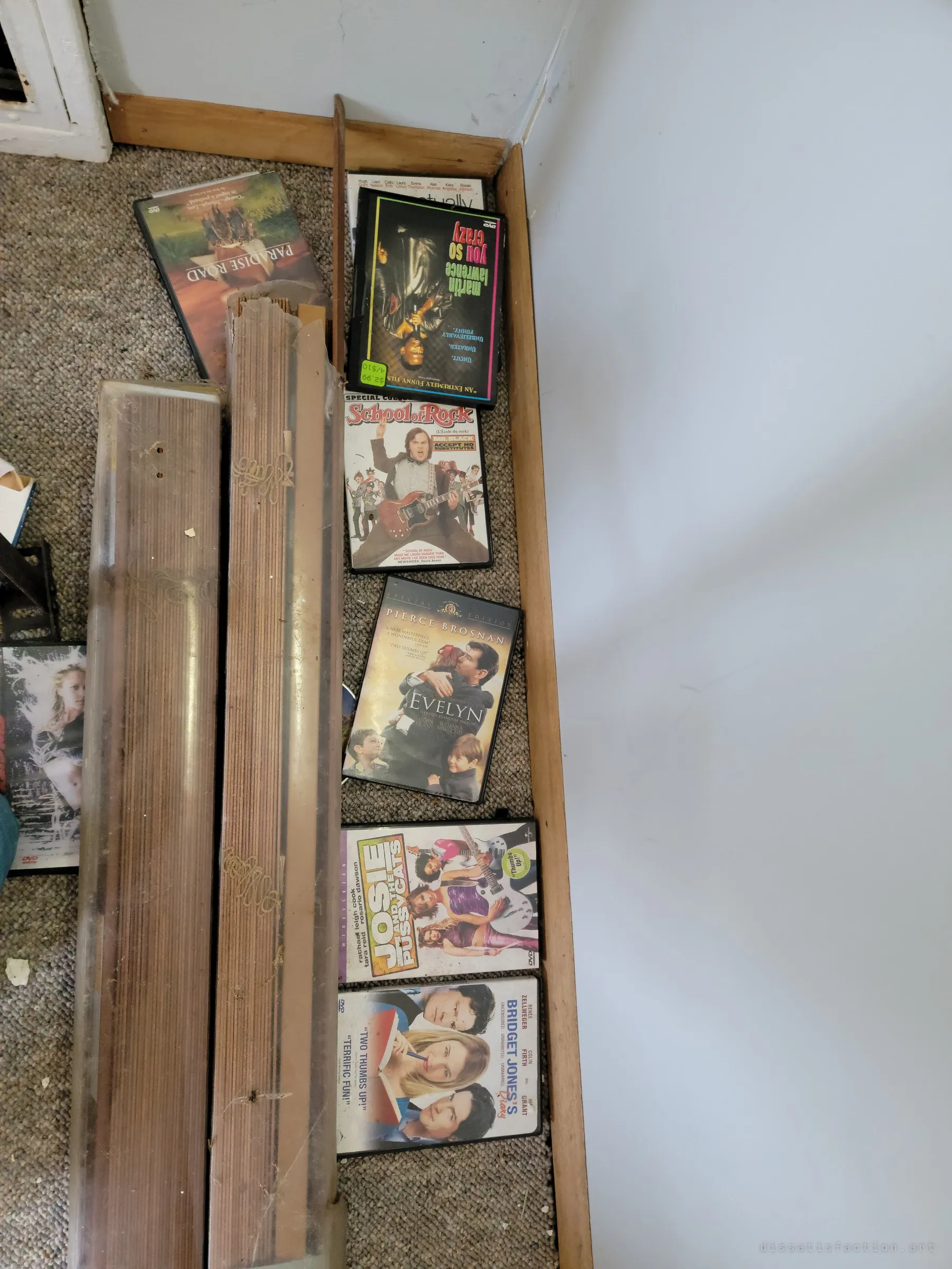 A row of old comedy and drama DVDs left on a carpeted floor beside rolled-up wooden blinds. Everyday leftovers of someone's movie nights.