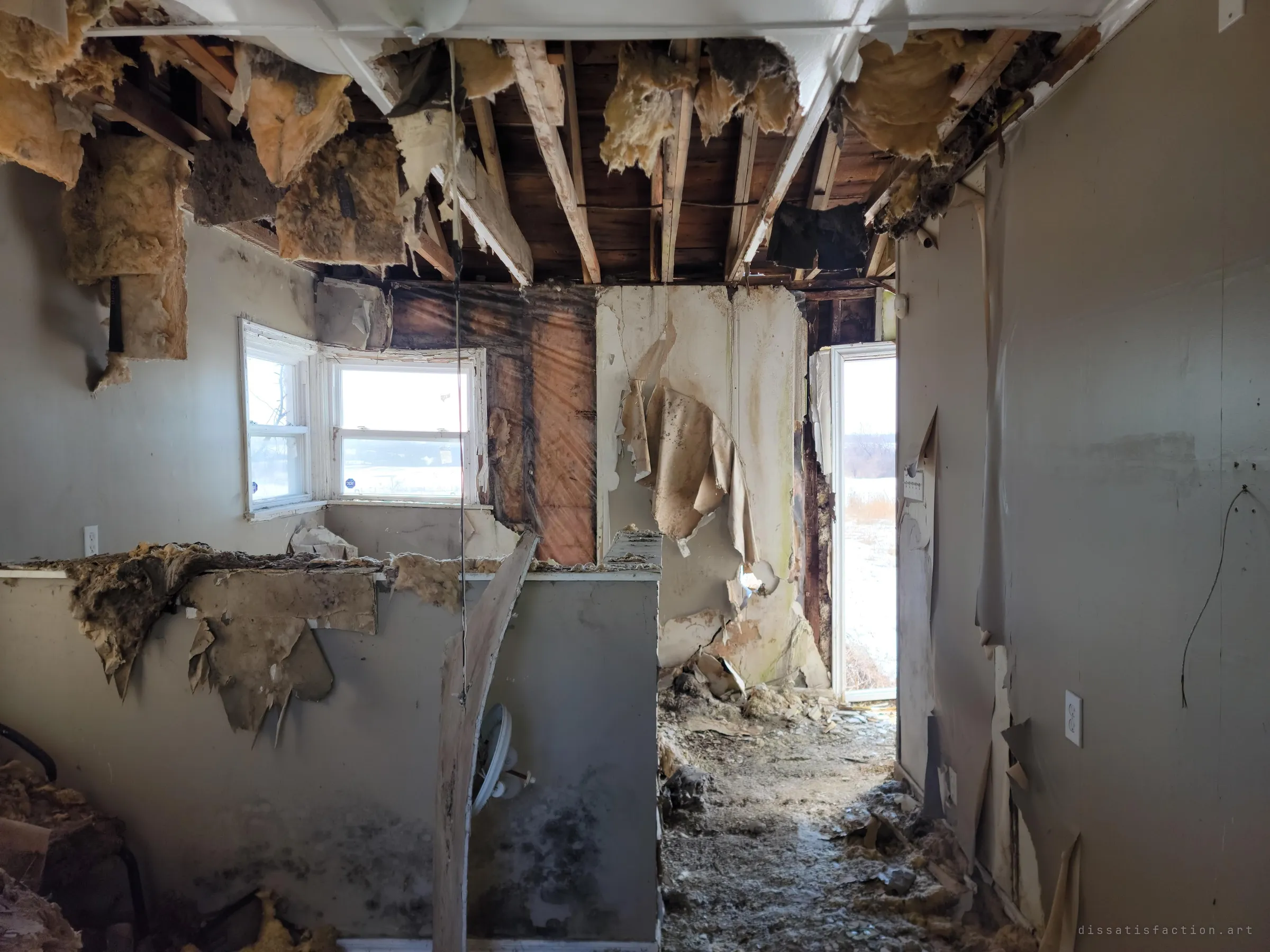 A gutted room with the ceiling torn open, insulation hanging from the joists and walls peeling away. An open doorway and windows show snowy fields outside.