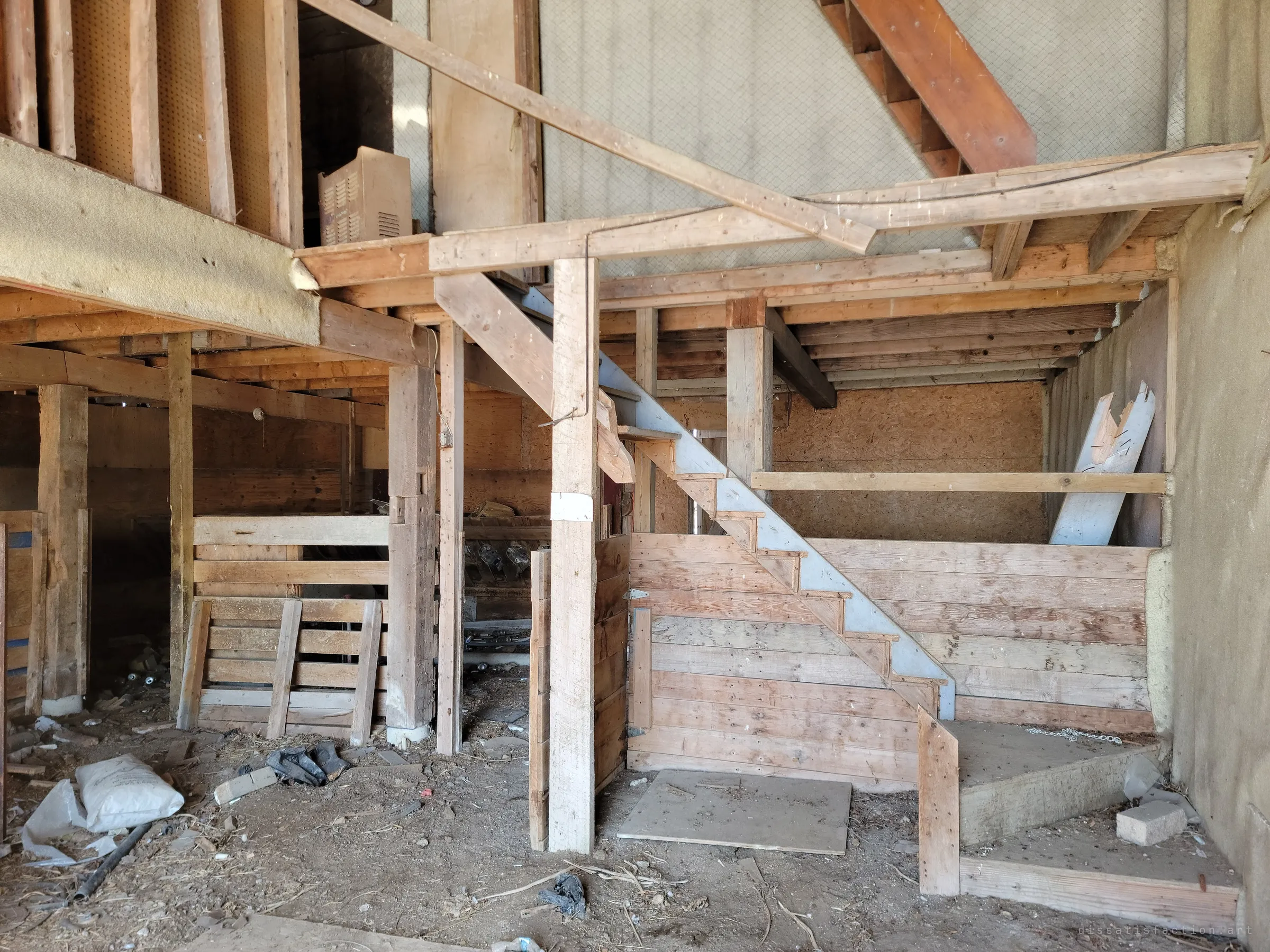 Inside a barn, a rough timber staircase climbs to a loft above wooden stalls and a dirt floor. The home-built framing shows years of patchwork repairs.