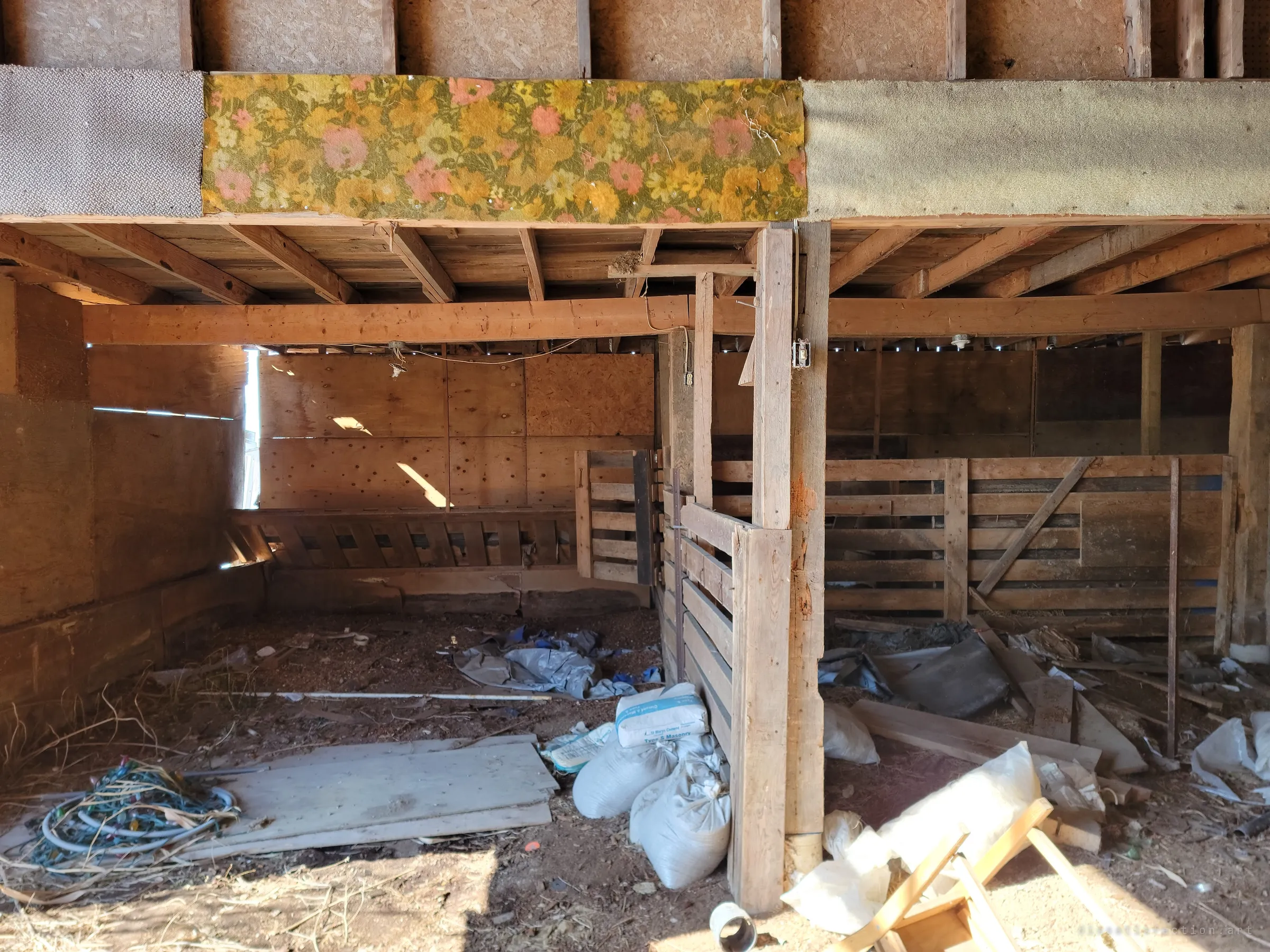 Empty animal pens under a loft edged with a strip of floral carpet, with sacks and scrap on the dirt floor. Sunlight leaks through gaps in the plywood walls.