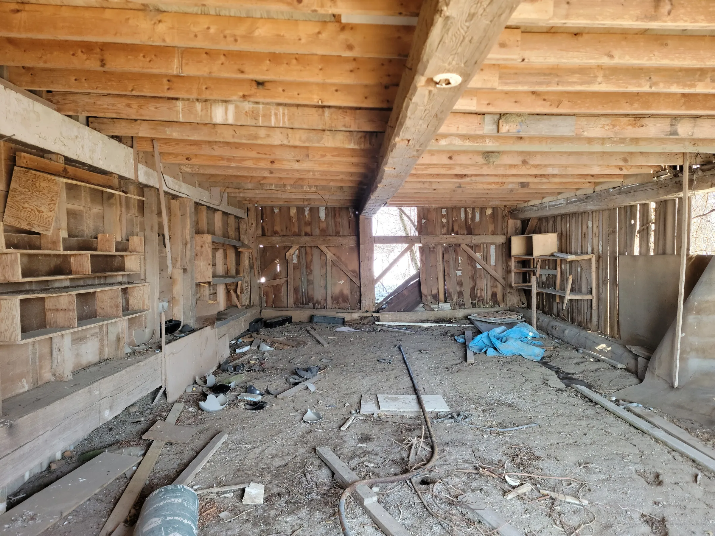 The low-ceilinged ground floor of an old barn, with a heavy central beam, empty shelves and broken pots scattered across the dirt. Daylight breaks through the weathered back wall.