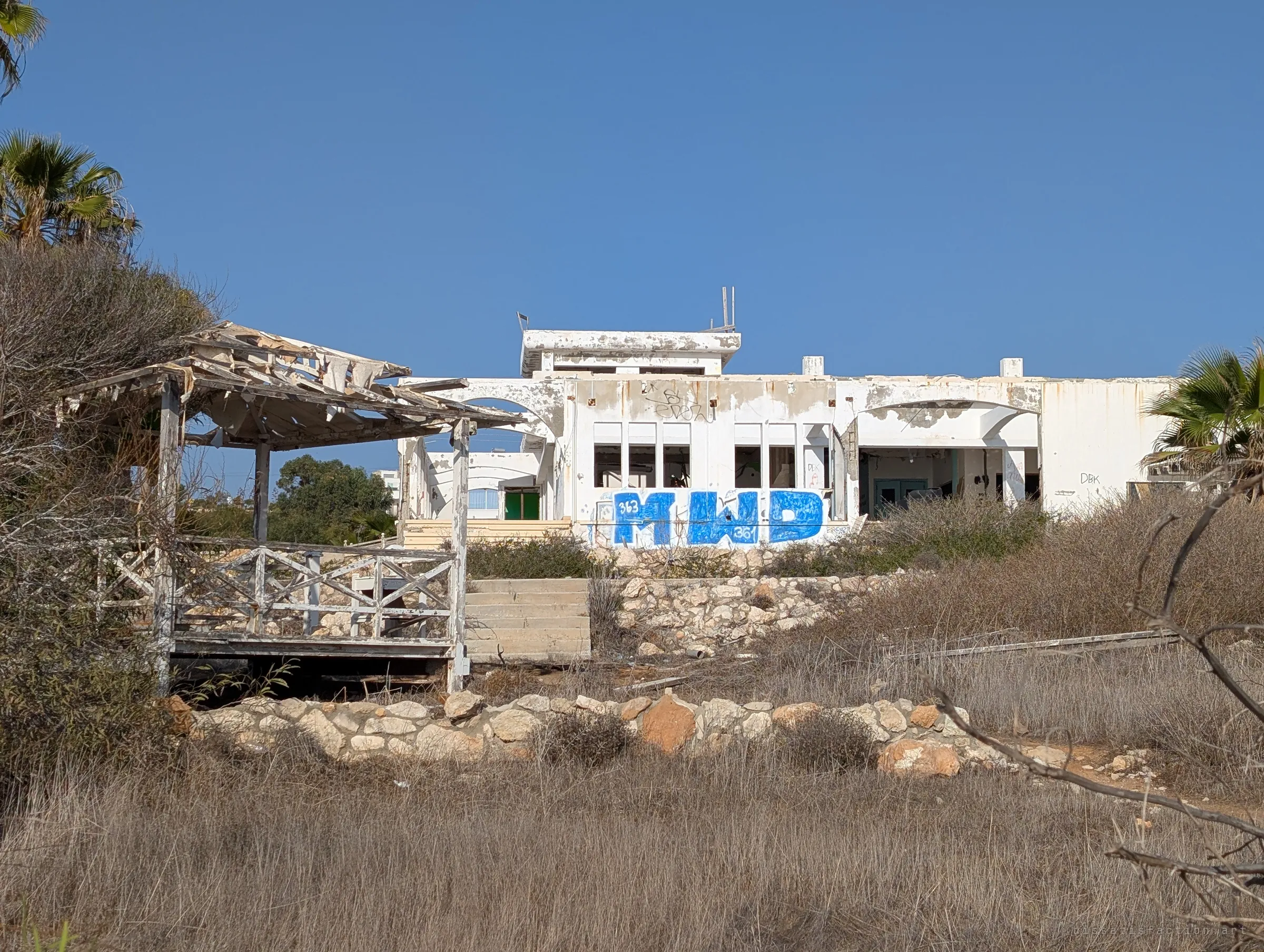 A collapsing wooden pergola and dry scrub lead up to a white, arcaded seaside building covered in a huge blue graffiti piece. It sets the scene: a sun-bleached resort left to the weeds.