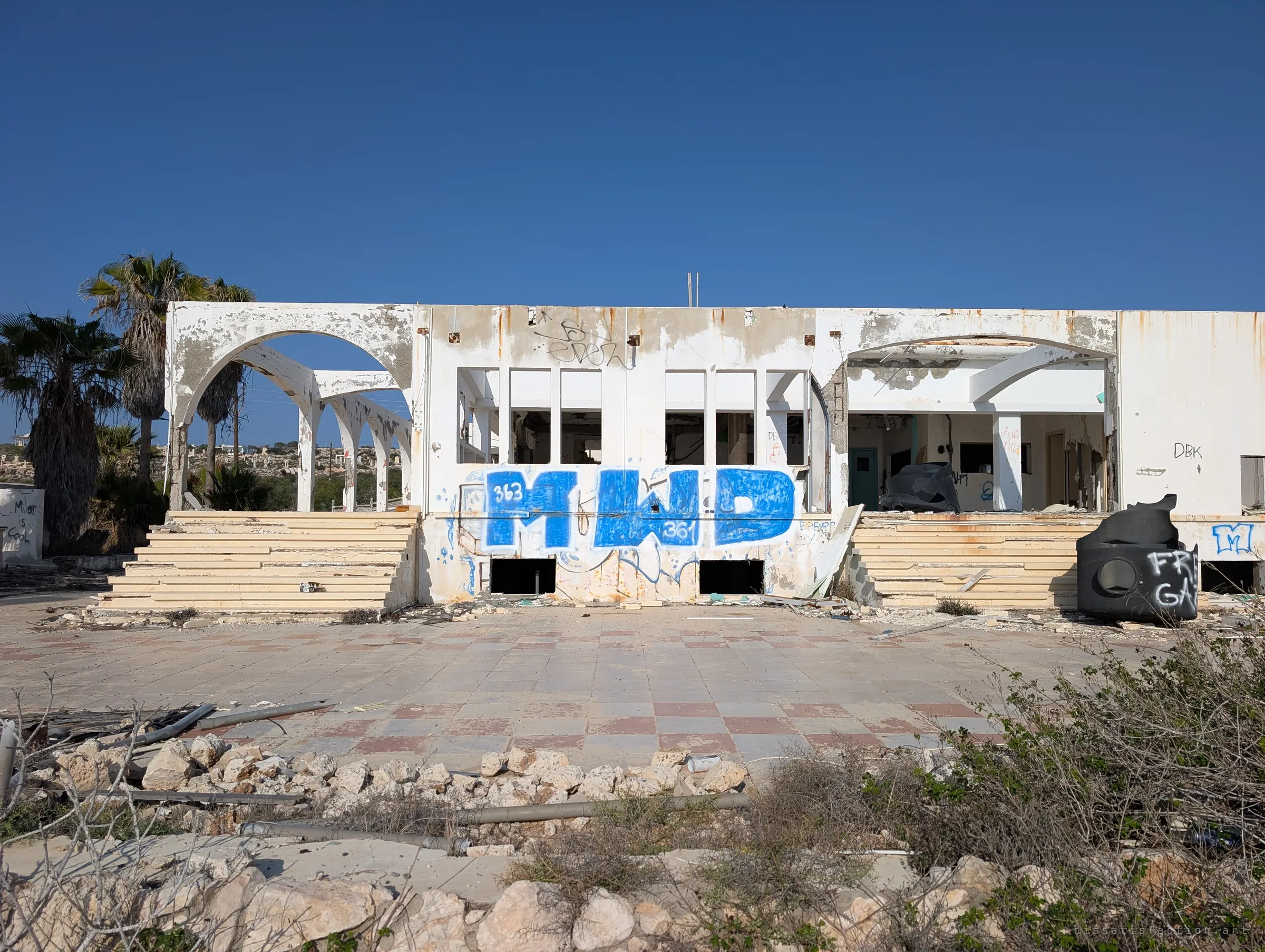 The front of the abandoned building seen square on across a cracked chequered terrace, broad steps rising to empty window frames under a giant blue tag. A black water tank lies tipped over at the right.