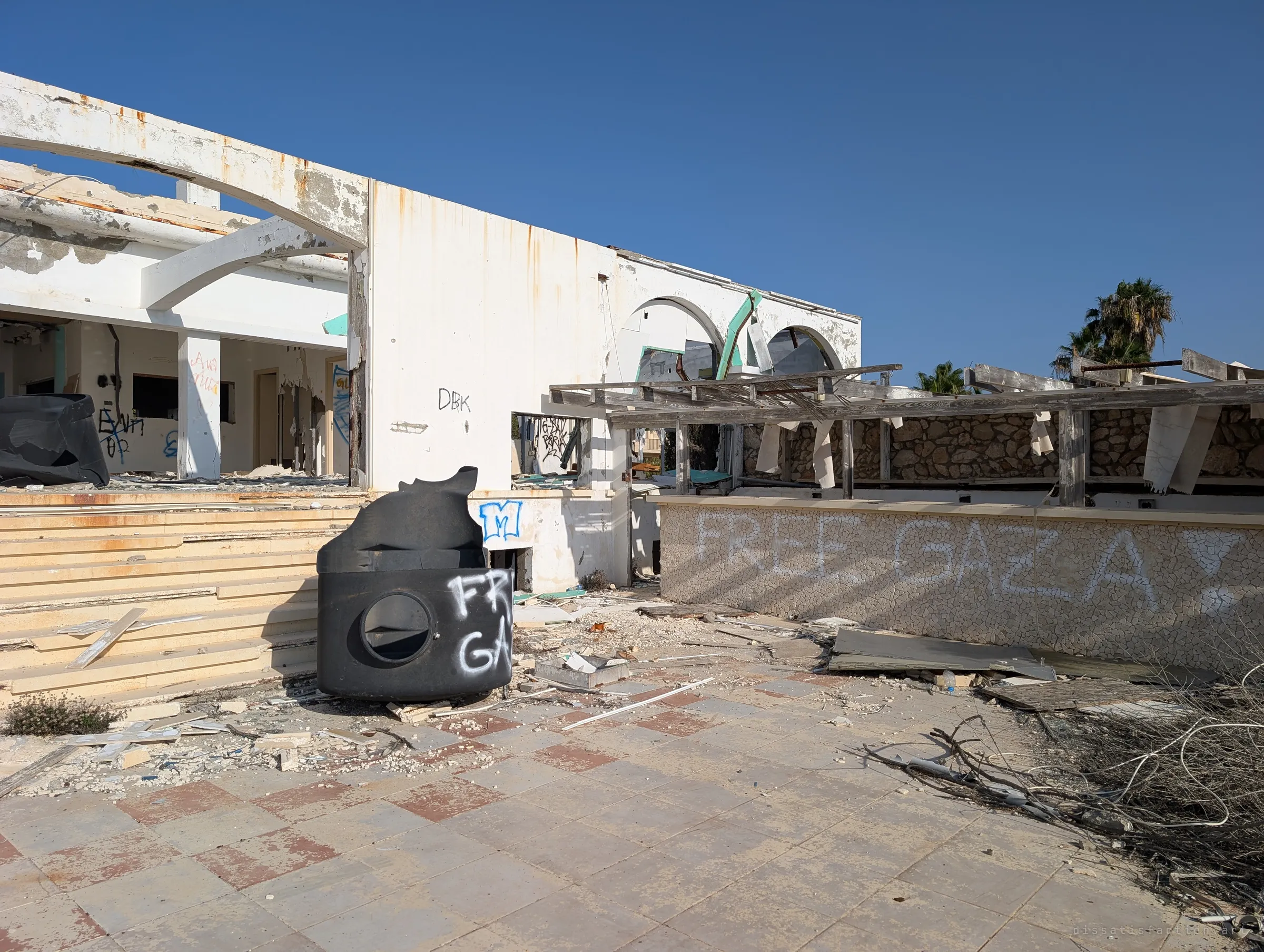 Side of the terrace with a toppled black water tank, a stripped pergola frame and a mosaic bar counter sprayed with a political slogan. Rubble and snapped boards cover the tiles.