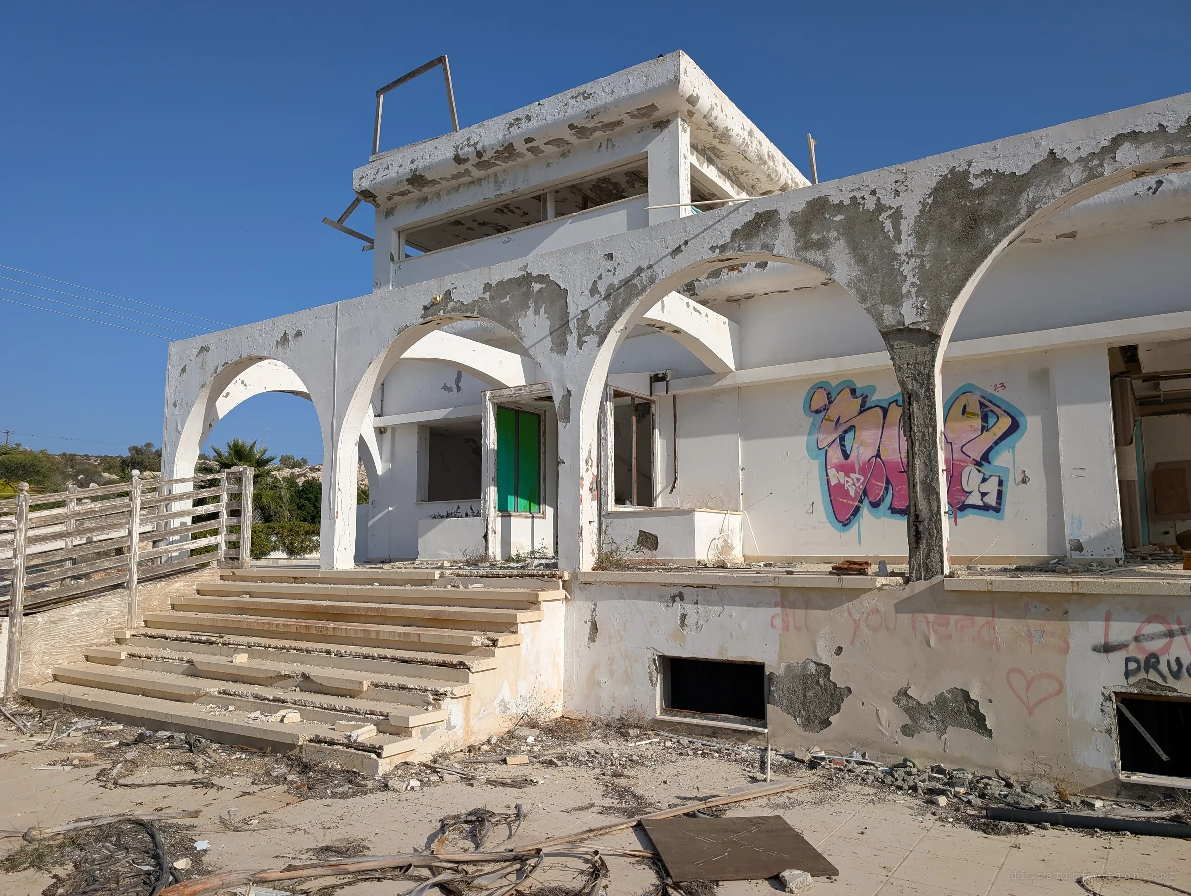 Arched colonnade and wide stairs beneath a small rooftop lookout, the plaster peeling in grey patches and a colourful graffiti piece on the inner wall. A green door still hangs in one opening.