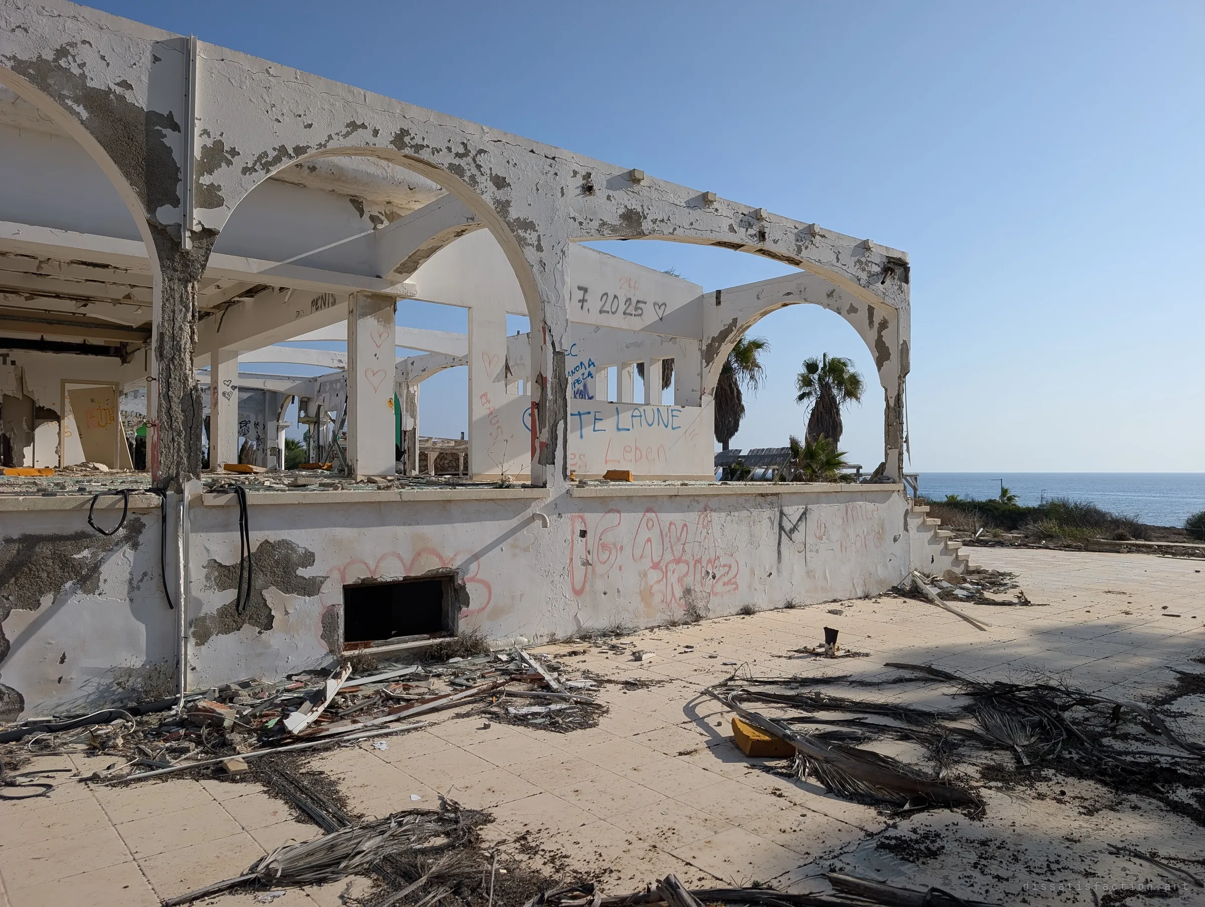 The open-sided pavilion from its seaward corner, arches framing palms and the blue sea beyond. Visitors' graffiti and dried palm fronds cover the walls and terrace.