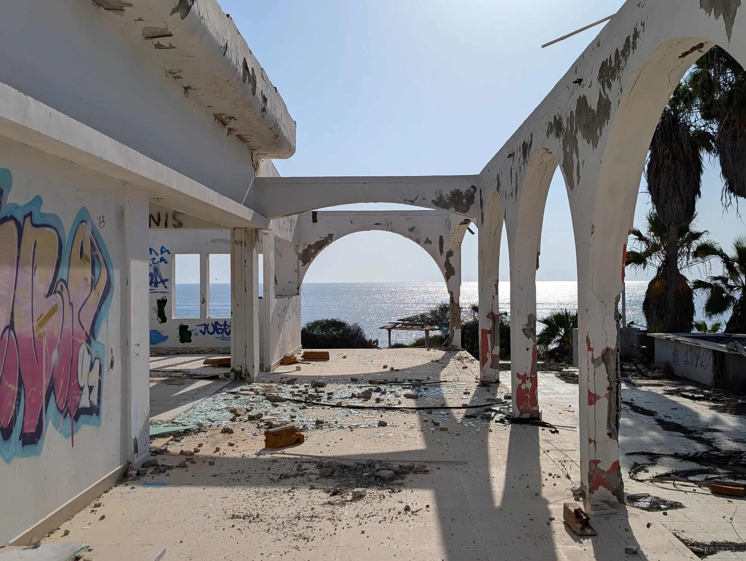 A roofless arcade runs out toward the glittering sea, its floor littered with shattered glass and chunks of masonry. Long shadows of the arches stripe the tiles.