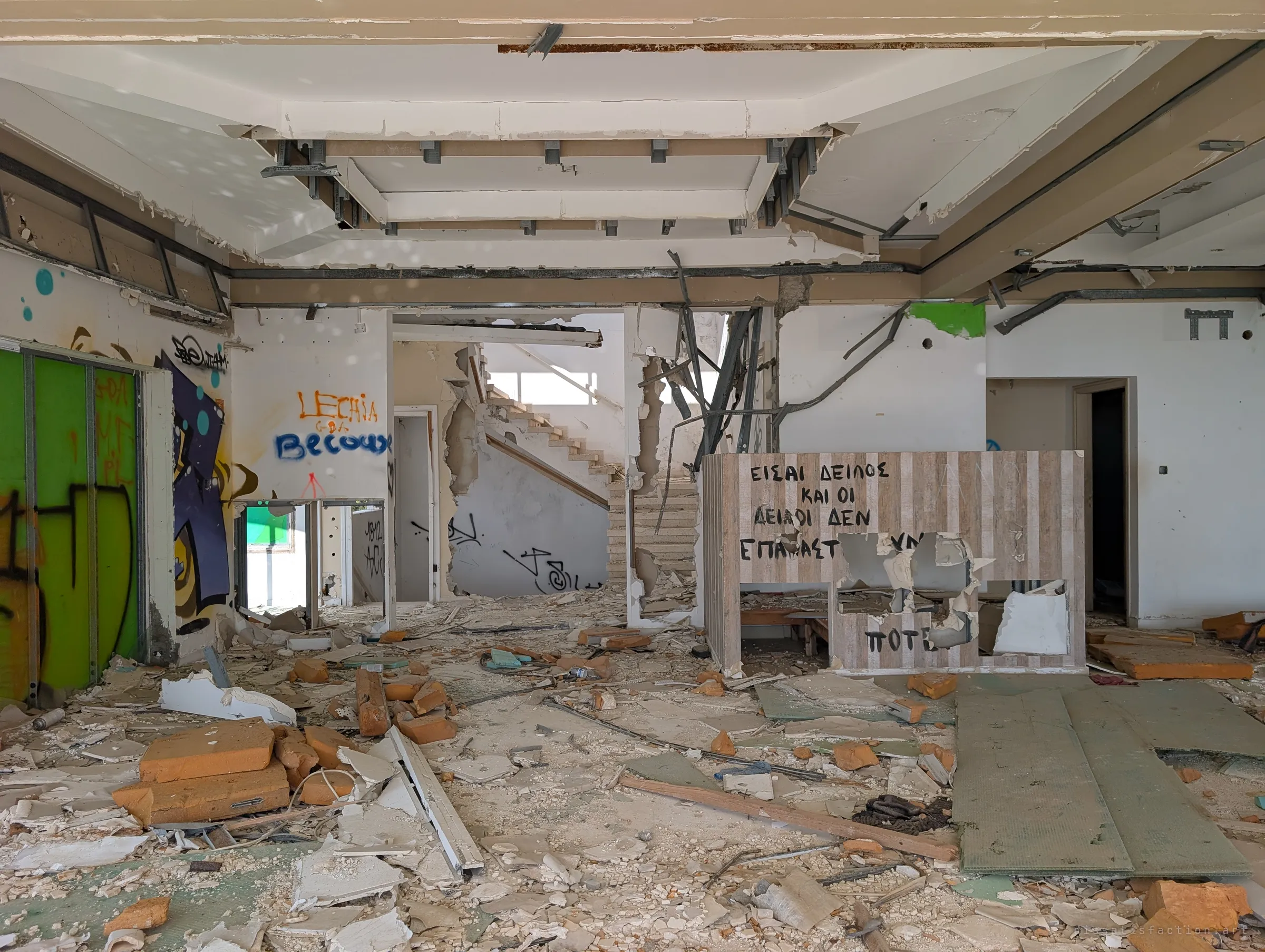 Inside the gutted main hall: ceiling panels hang open, a stair climbs through a smashed wall and a counter carries hand-painted graffiti. Insulation and plasterboard are piled across the floor.