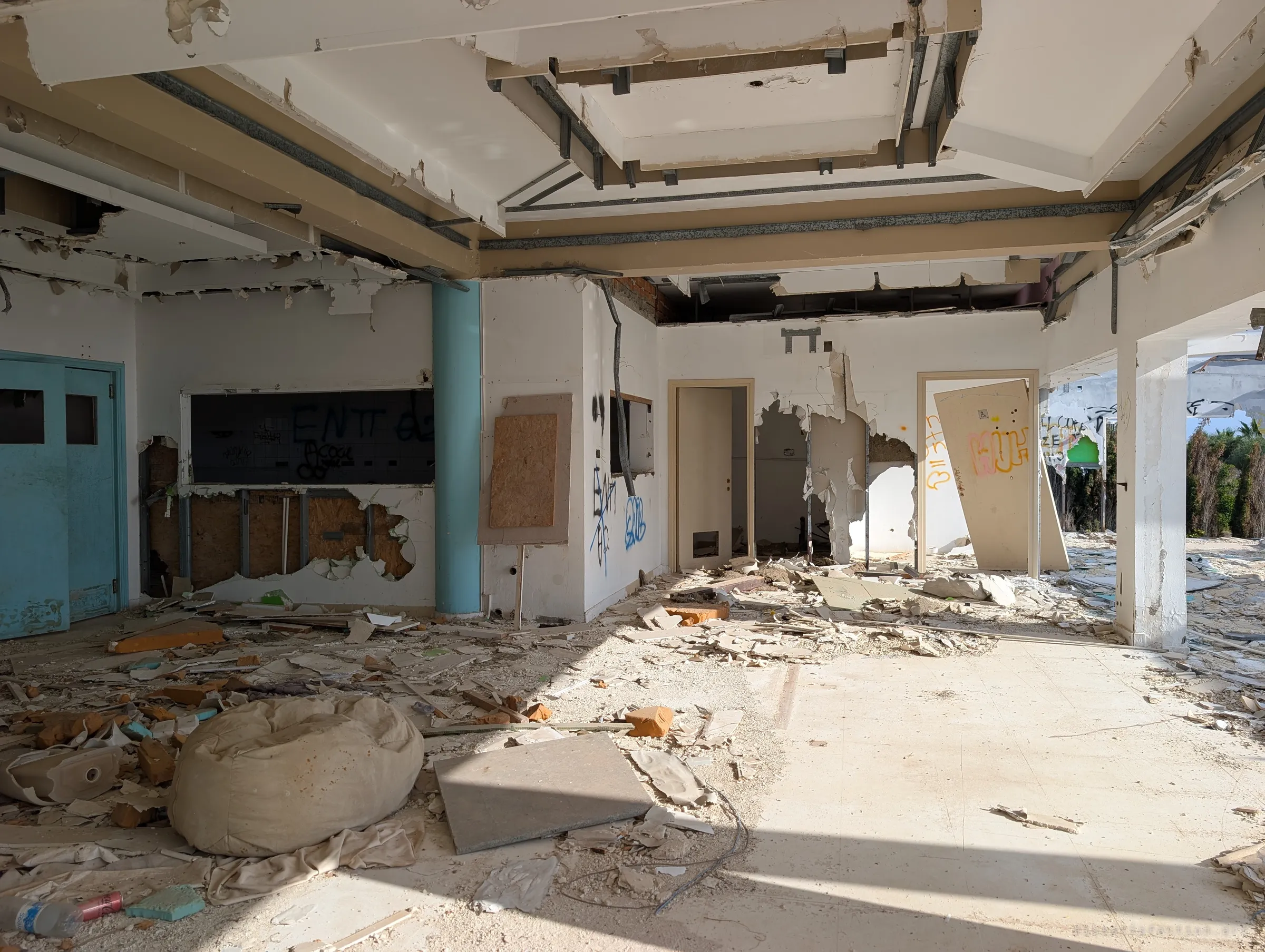 A sunlit interior room with a turquoise column, a tiled serving hatch and doors torn from their frames. A deflated bean bag sits in the debris, a small trace of the people who once lounged here.